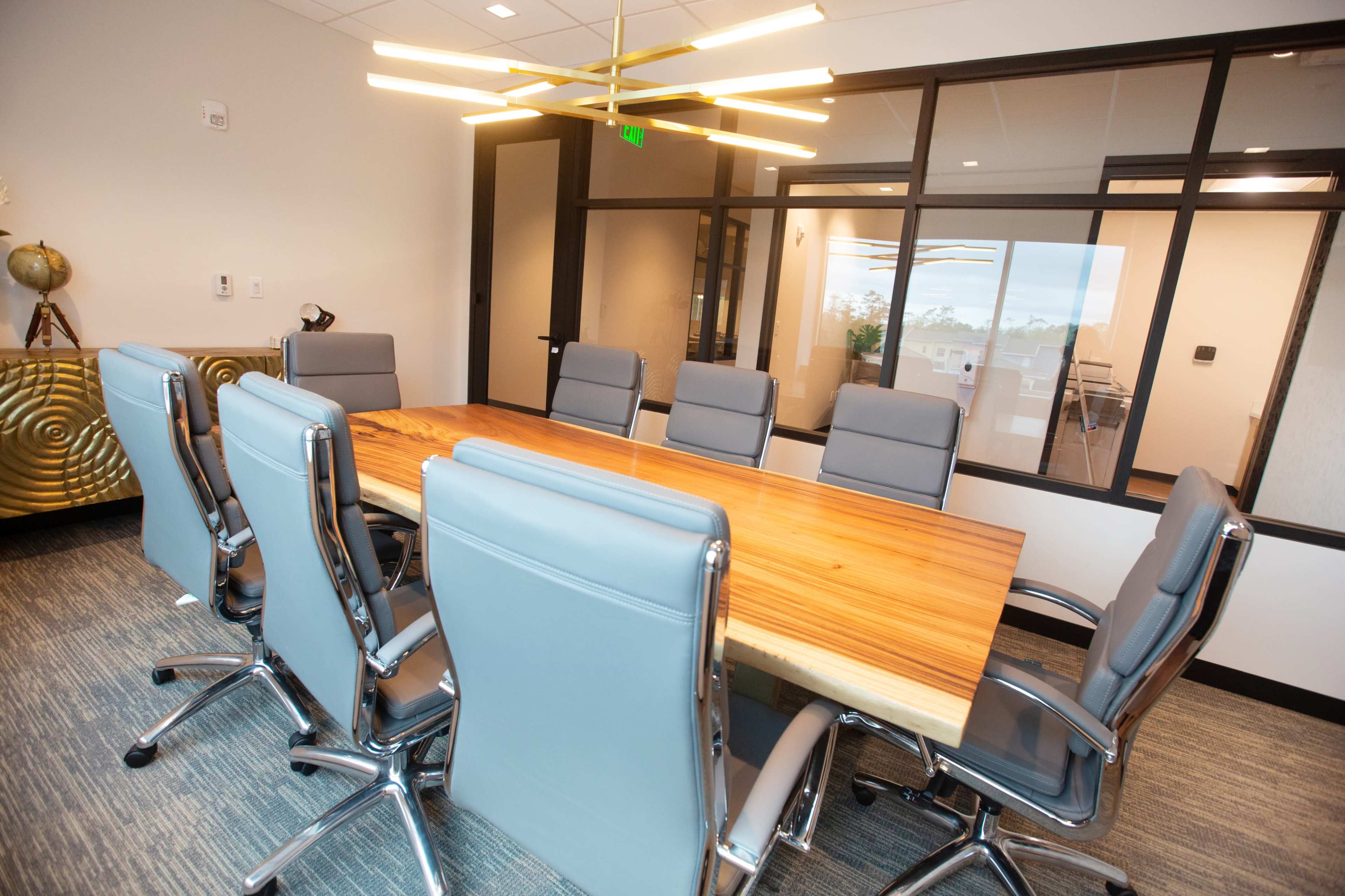 A modern conference room featuring a long wooden table surrounded by eight gray office chairs and illuminated by a contemporary light fixture.