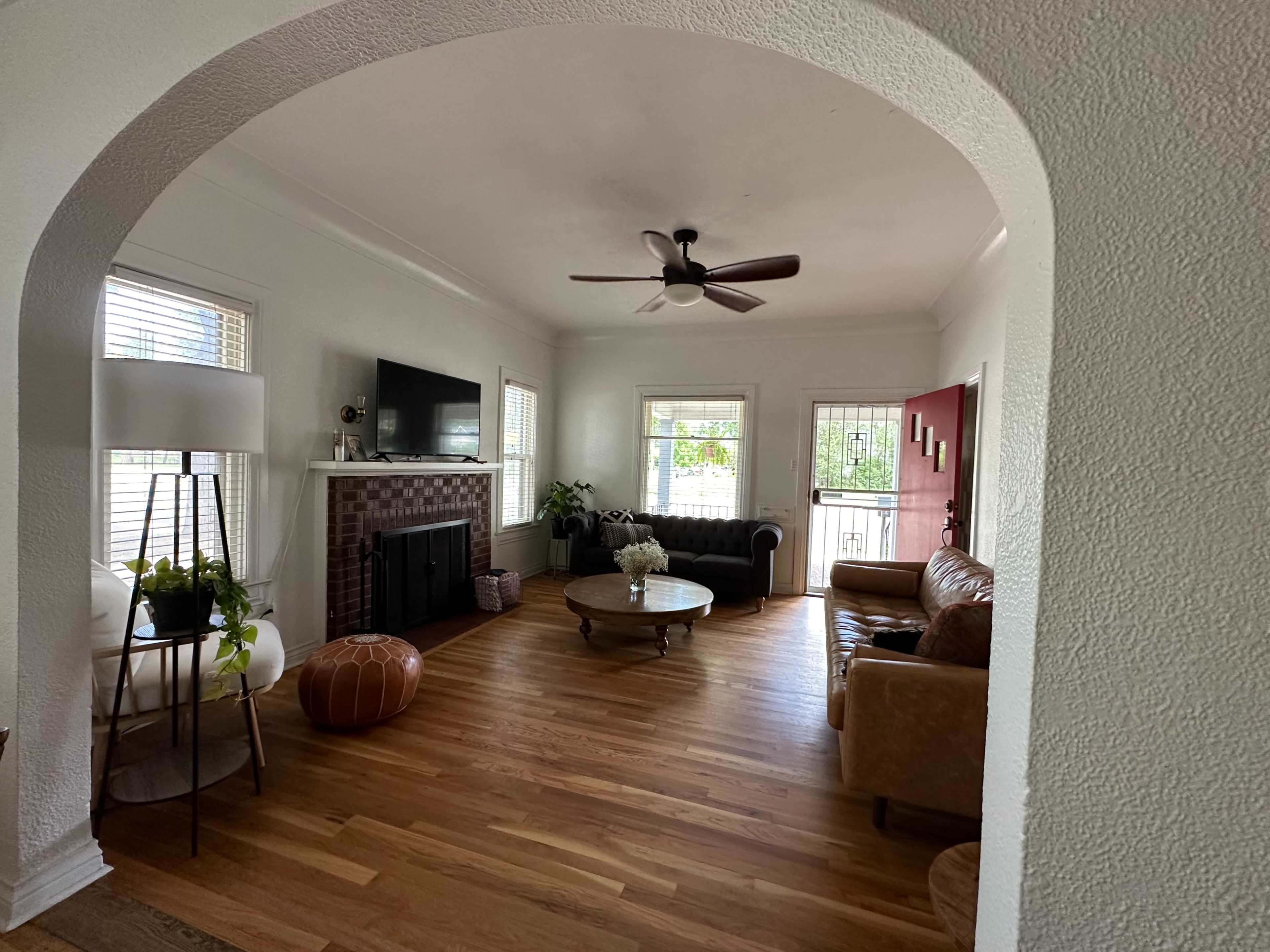 The image shows a bright living room featuring a leather couch, a coffee table, a TV mounted on the wall, and large windows with natural light.
