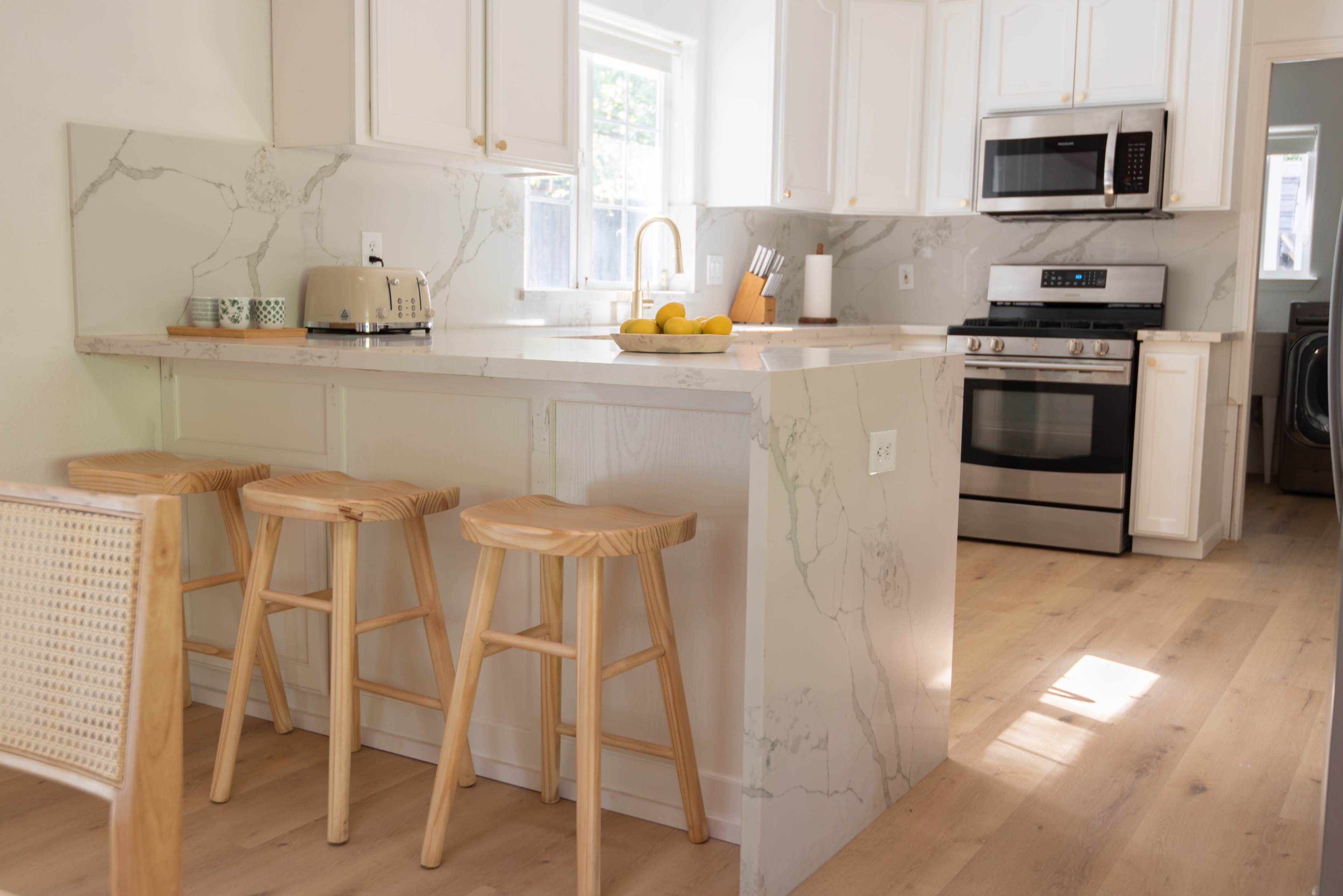 A modern kitchen features white cabinetry, a marble countertop island with three wooden stools, and stainless steel appliances.
