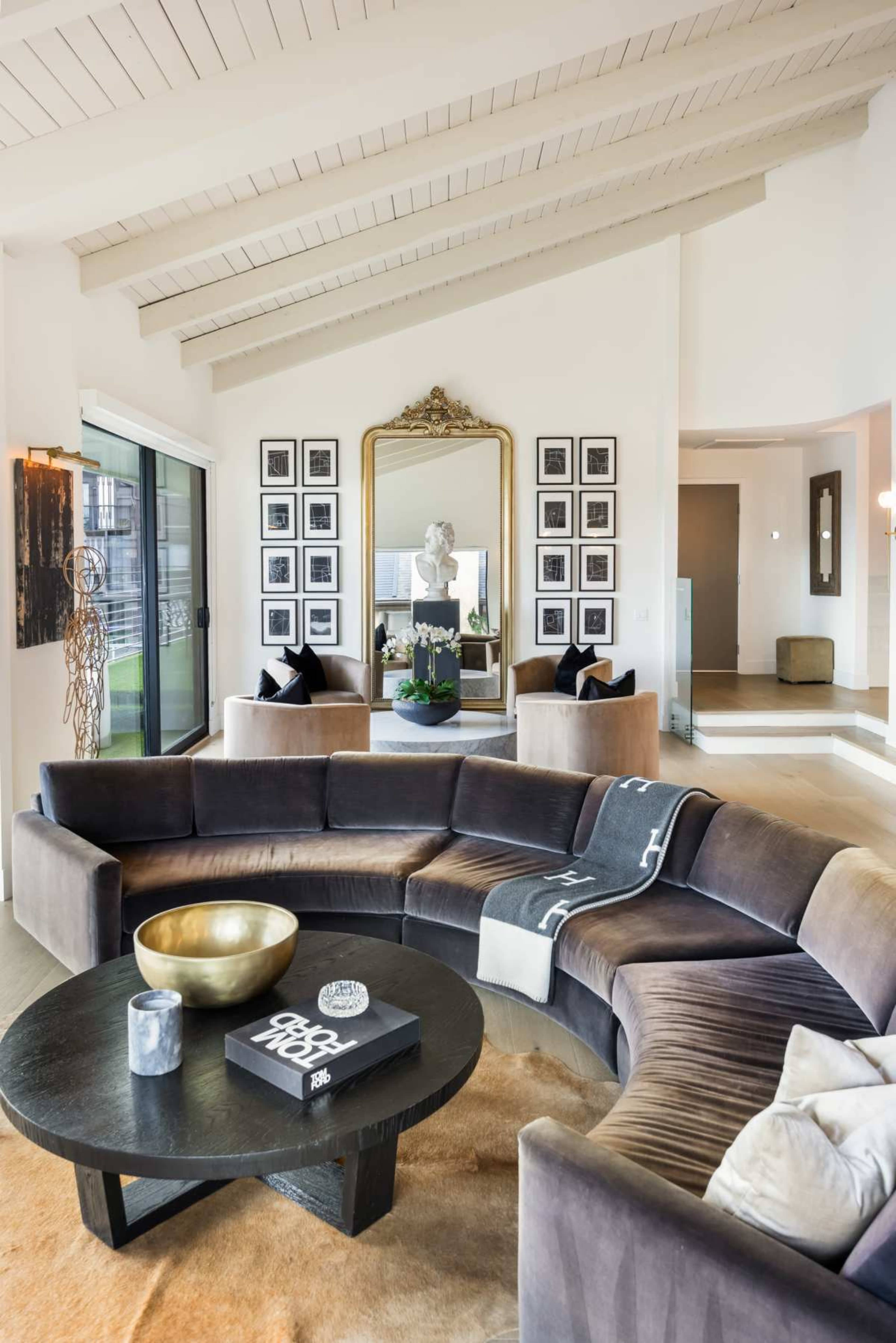 A modern living room features a curved sectional sofa, a round coffee table, a large mirror, and a gallery wall of black and white photos.