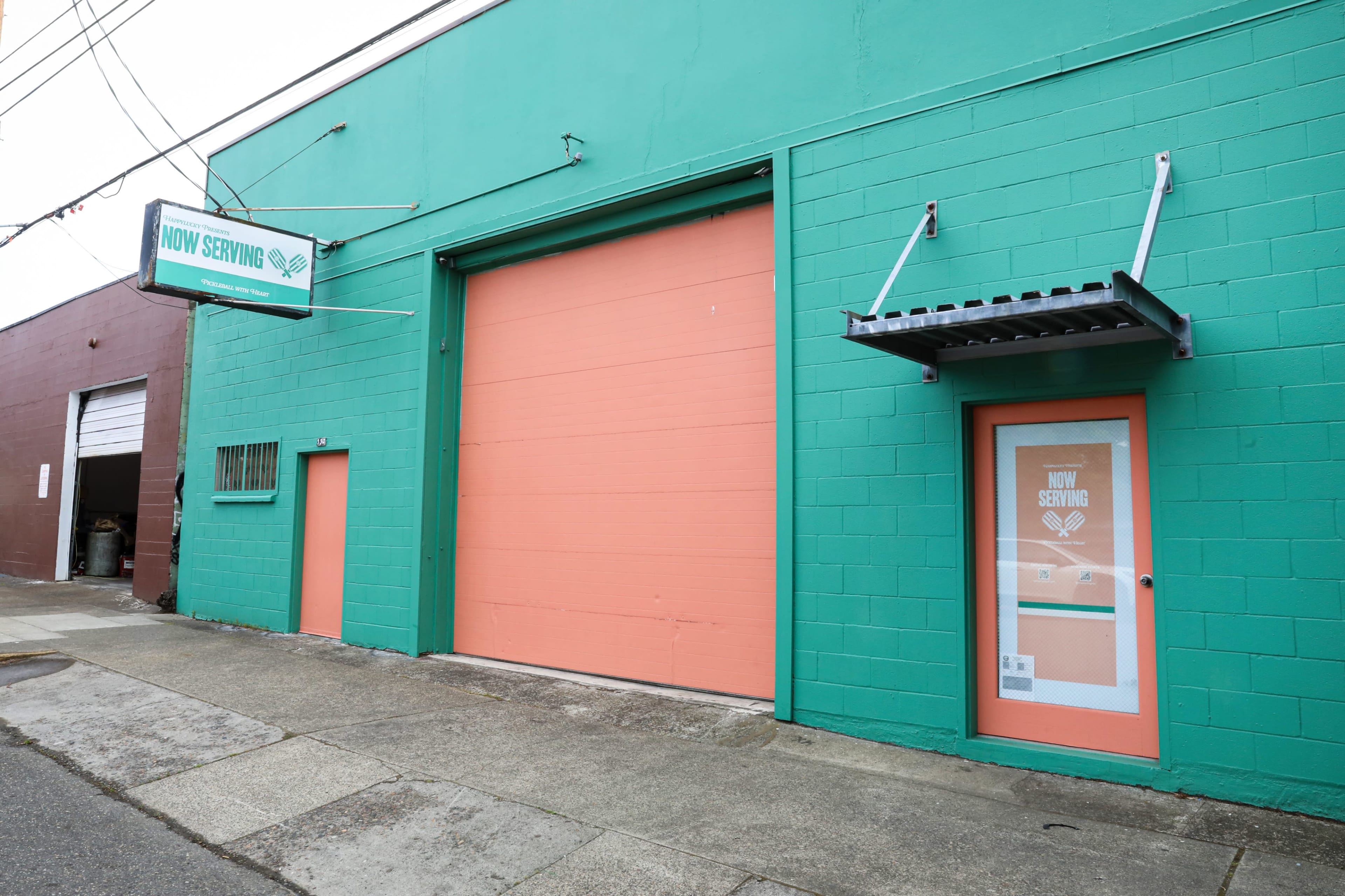 The image shows a brightly colored building with a turquoise exterior and a large pink garage door, featuring a smaller door beside it with a sign that reads "NOW SERVING."