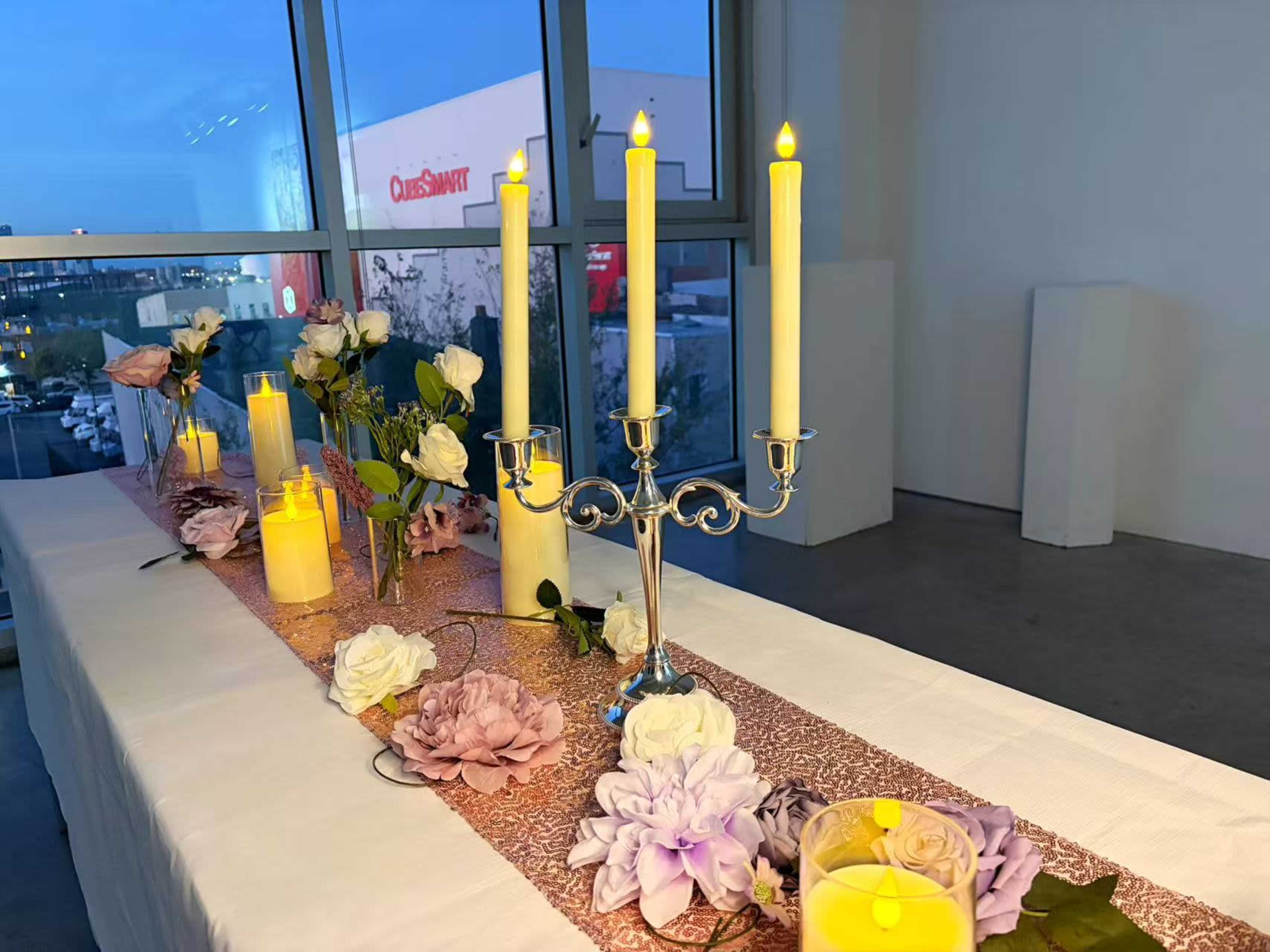 A table decorated with flowers and candles is set up in a bright room with large windows, revealing an outdoor view.