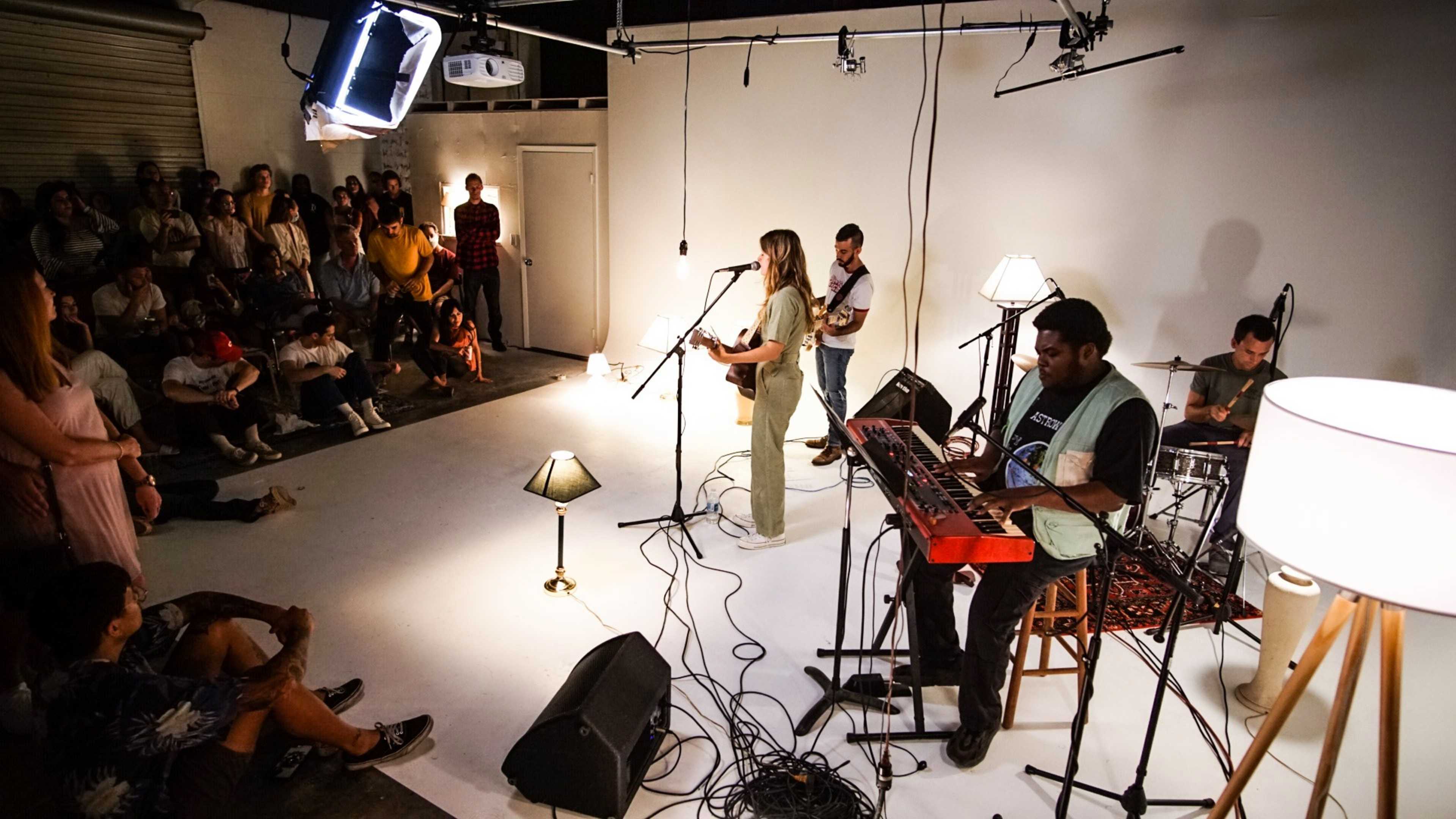 A band performs on a minimalistic stage in front of an audience seated on the floor.