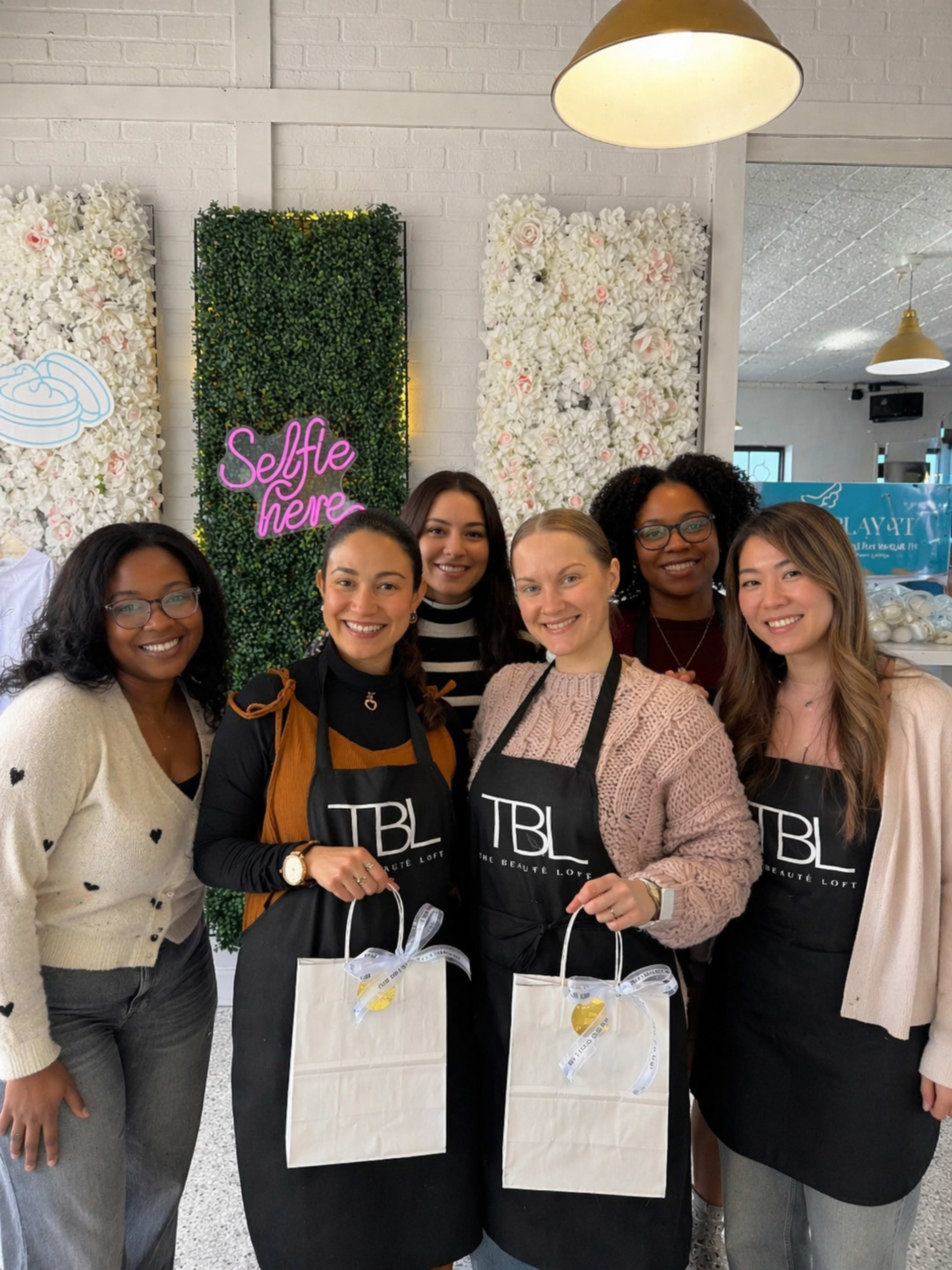 A group of six women, all wearing black aprons with "TBL" printed on them, stands together in front of a decorative wall featuring flowers and a neon sign that says "Selfie here."