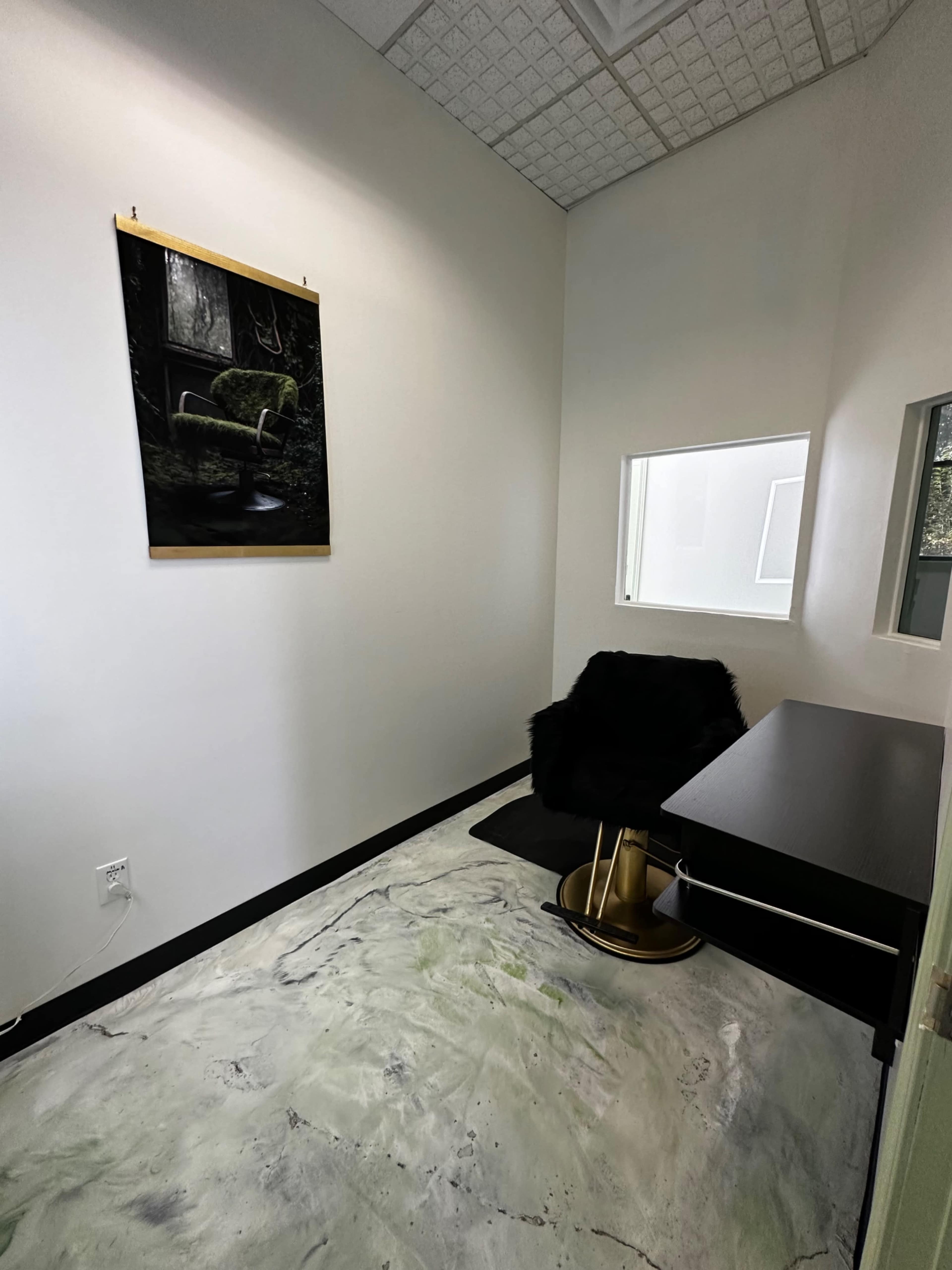 The image shows a small, modern room featuring a black chair, a sleek desk, and a wall-mounted artwork, with a window allowing natural light.
