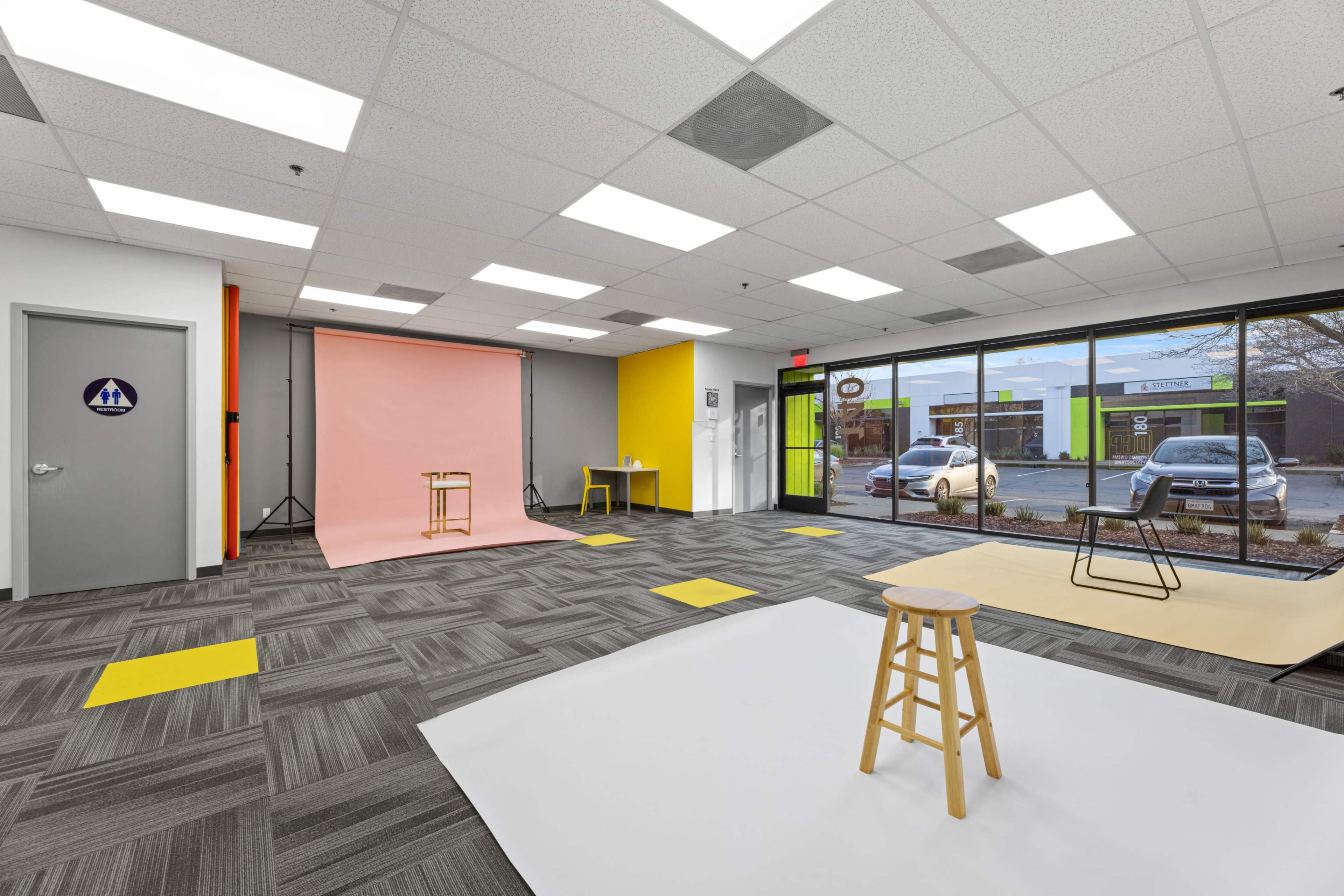 The image shows a spacious studio with gray carpeting, a large window, a pink backdrop, a yellow accent wall, and various props including stools and a table.