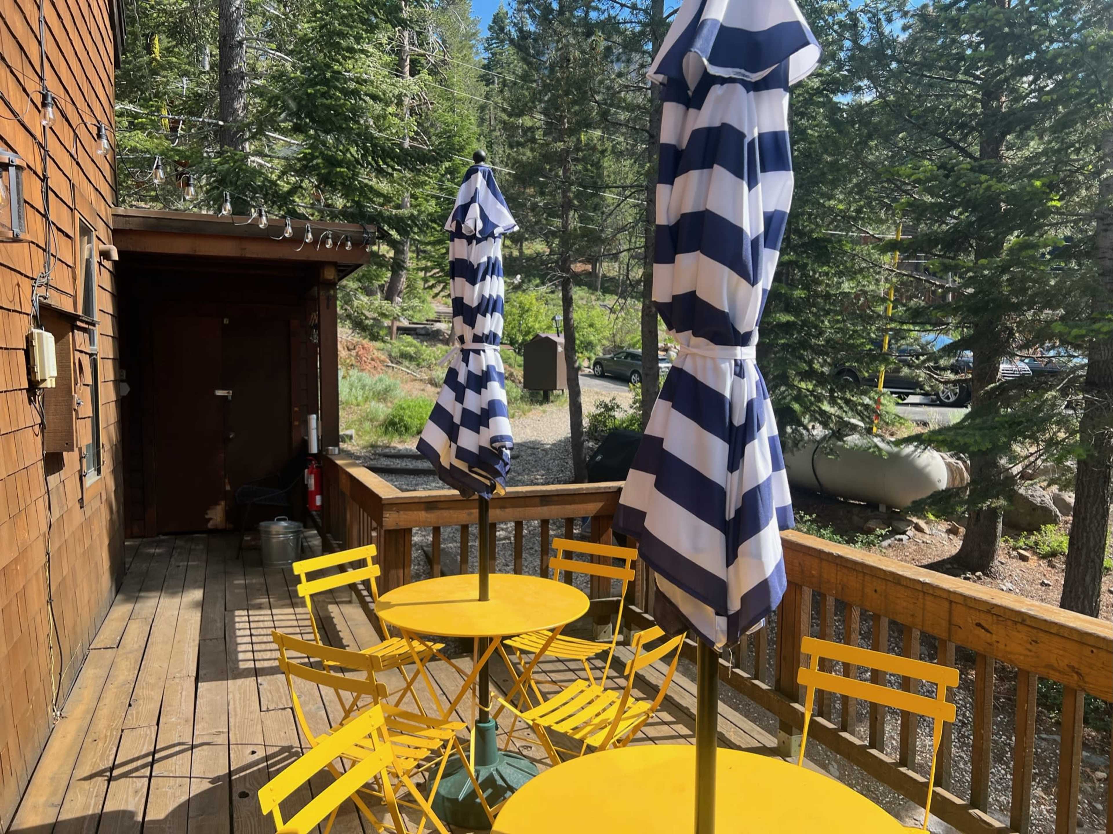 A wooden deck features yellow tables and blue-and-white striped umbrellas surrounded by trees.