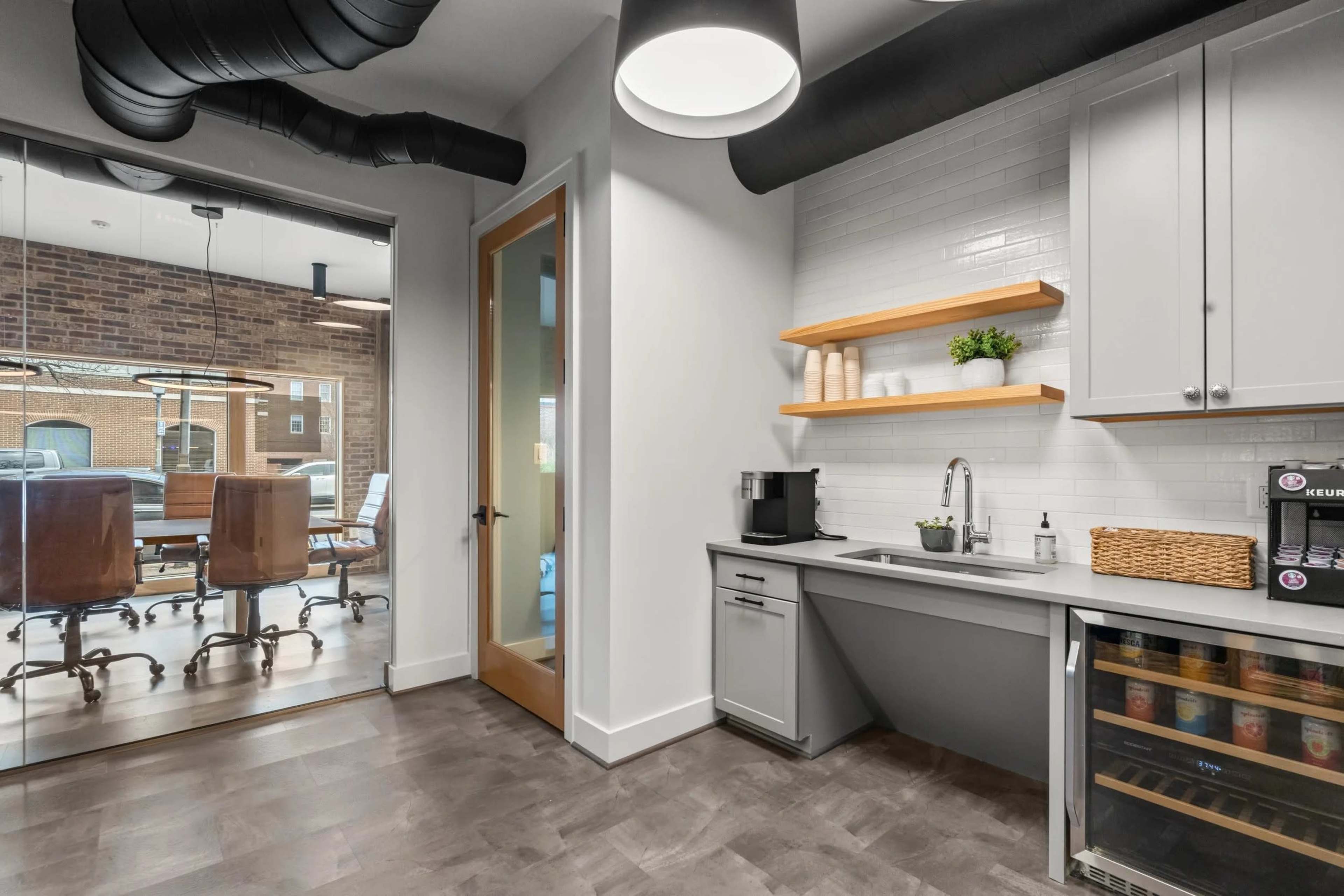 The image shows a modern office kitchenette with gray cabinetry, a sink, and a coffee maker, adjacent to a glass-walled conference room.