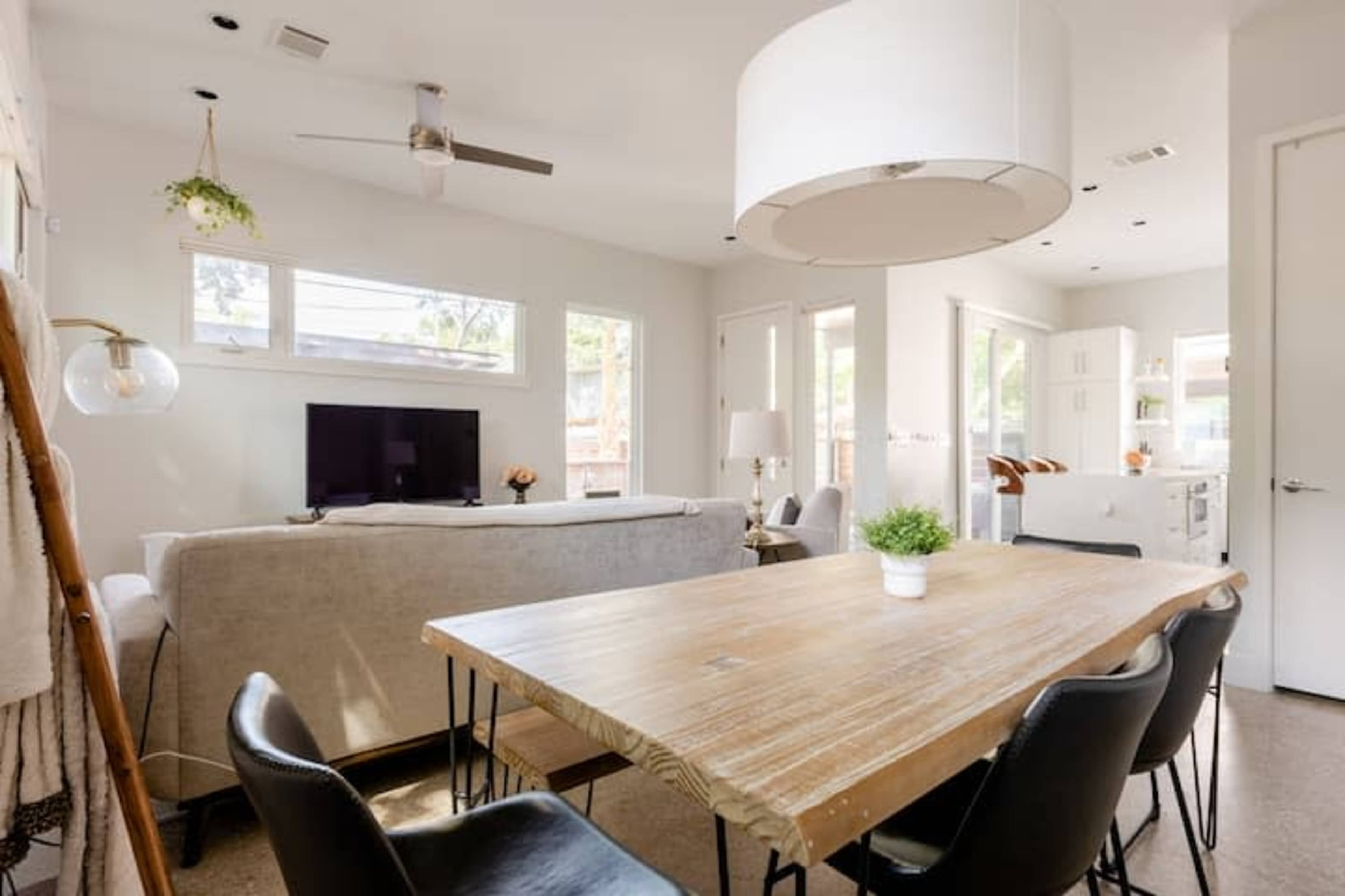 This image shows a modern living and dining area with a wooden dining table, black chairs, and a light-colored sofa facing a television.