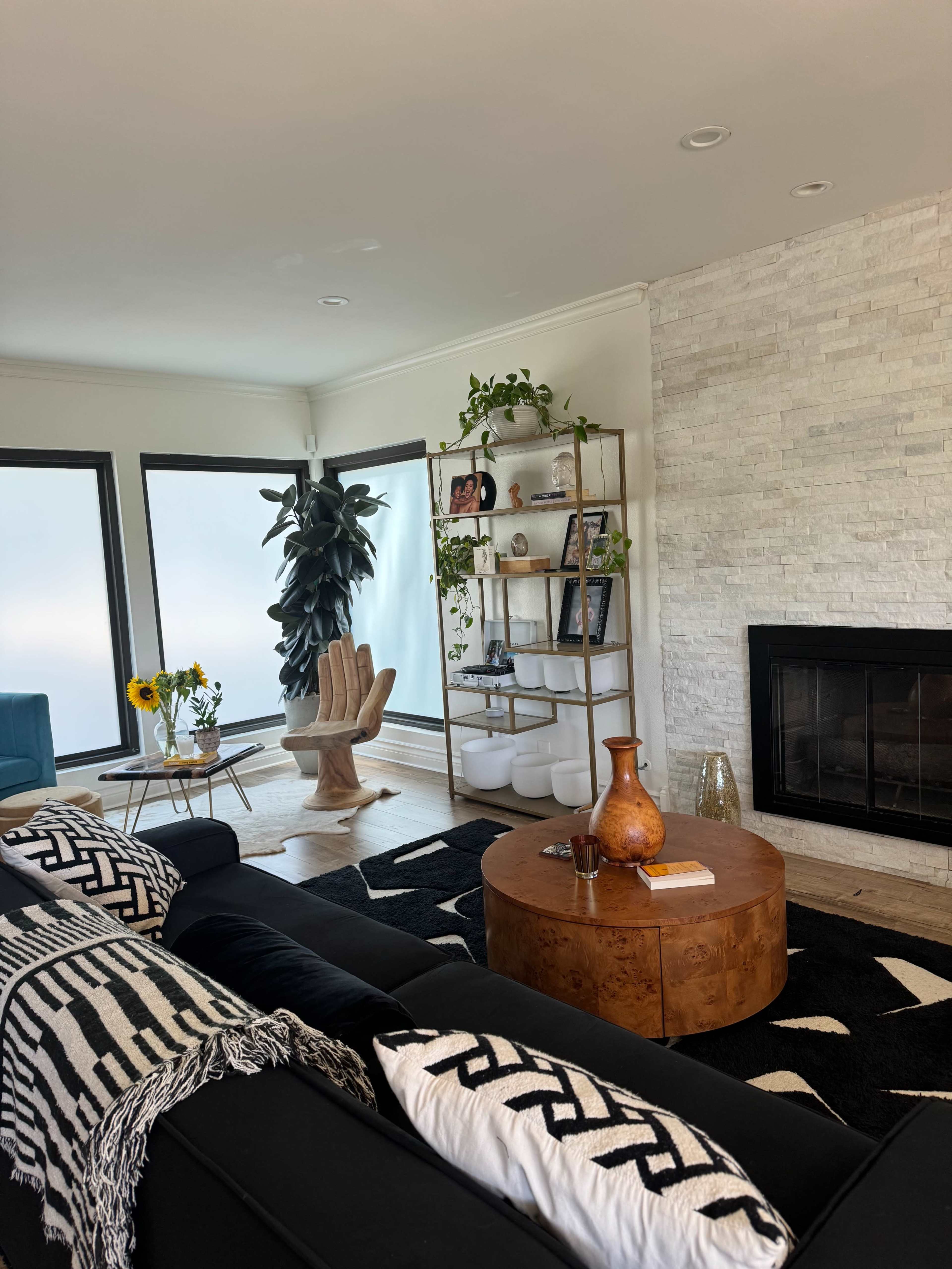 The living room features a black sofa, a round wooden coffee table, a bookshelf filled with decor, and large windows that allow natural light to fill the space.
