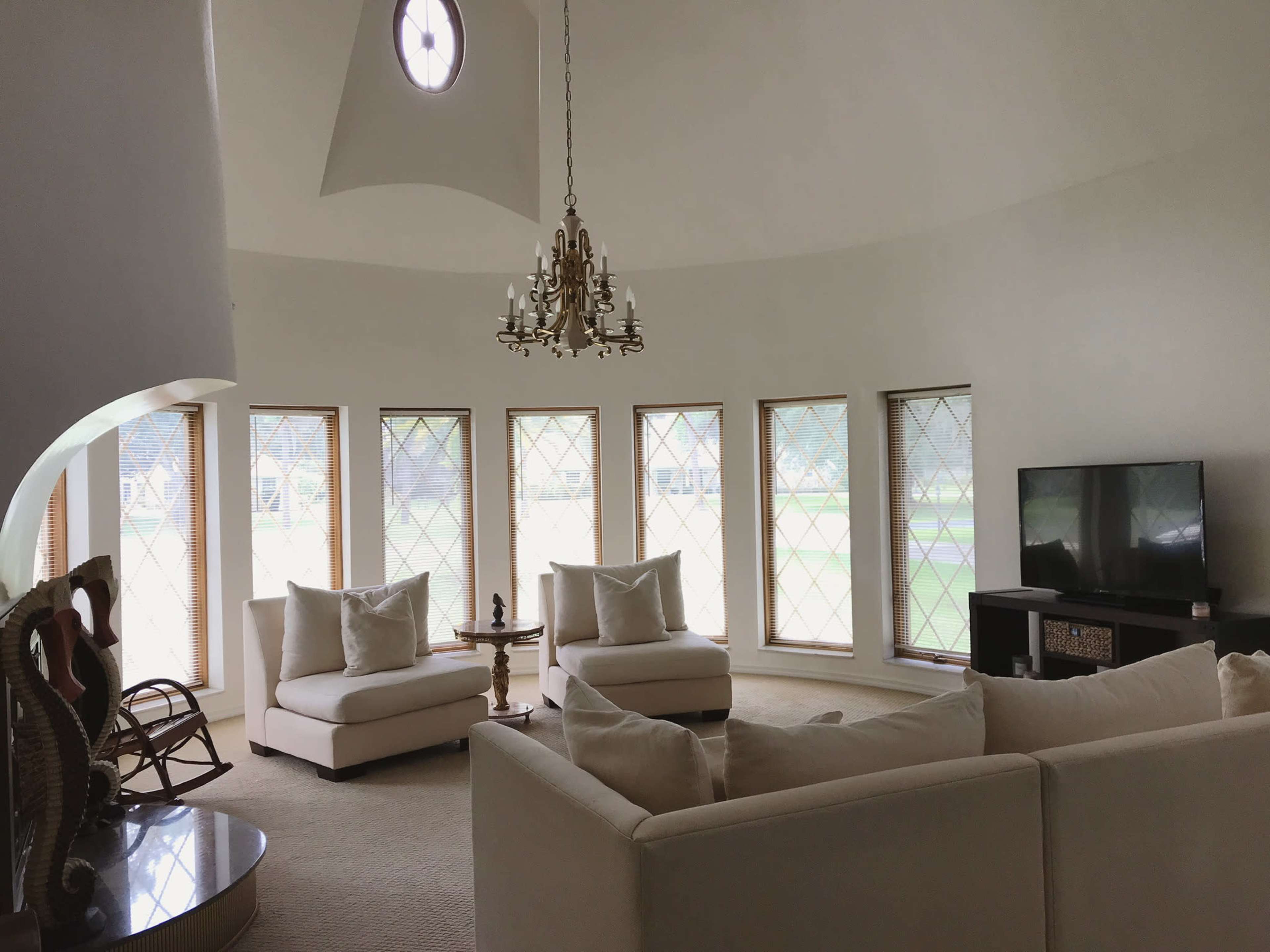 A circular living room features a chandelier, two armchairs, and a large sectional couch arranged around a coffee table, with tall windows providing natural light.
