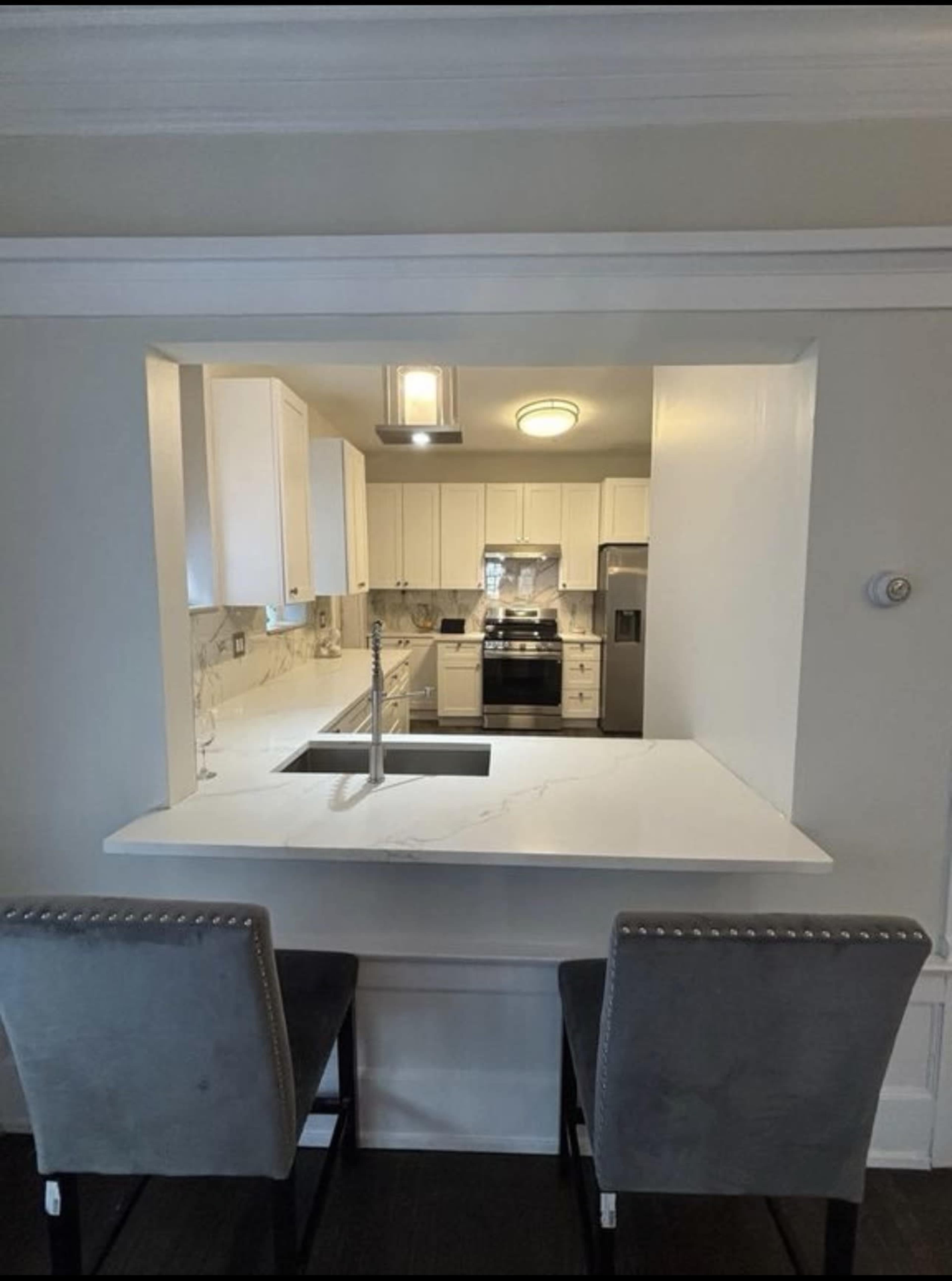 The image shows a modern kitchen with white cabinets, stainless steel appliances, and a marble countertop with two gray bar stools positioned in front of it.