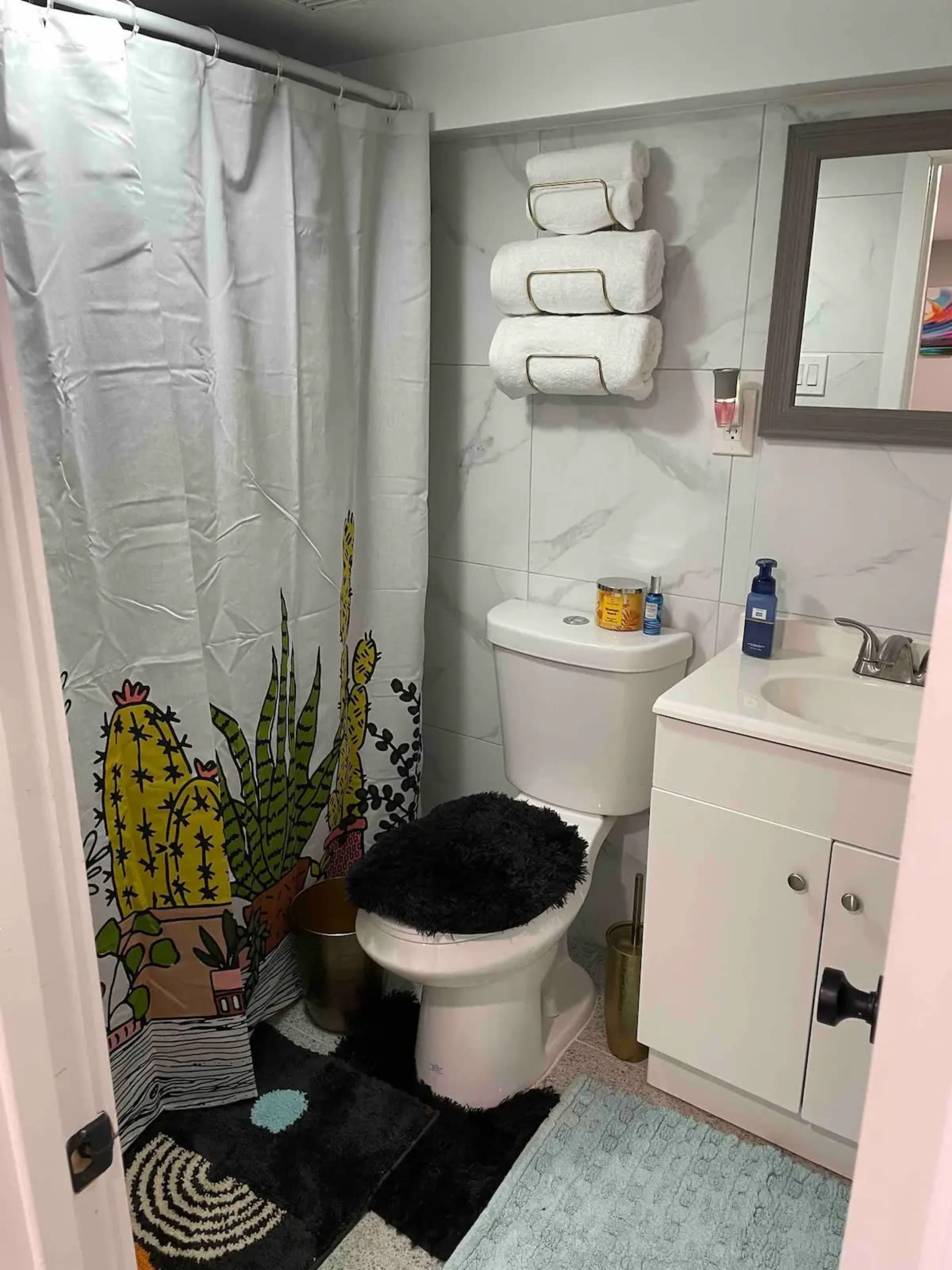 The bathroom features a toilet, a shower with a cactus-patterned curtain, stacked towels, and a sink with various toiletries.
