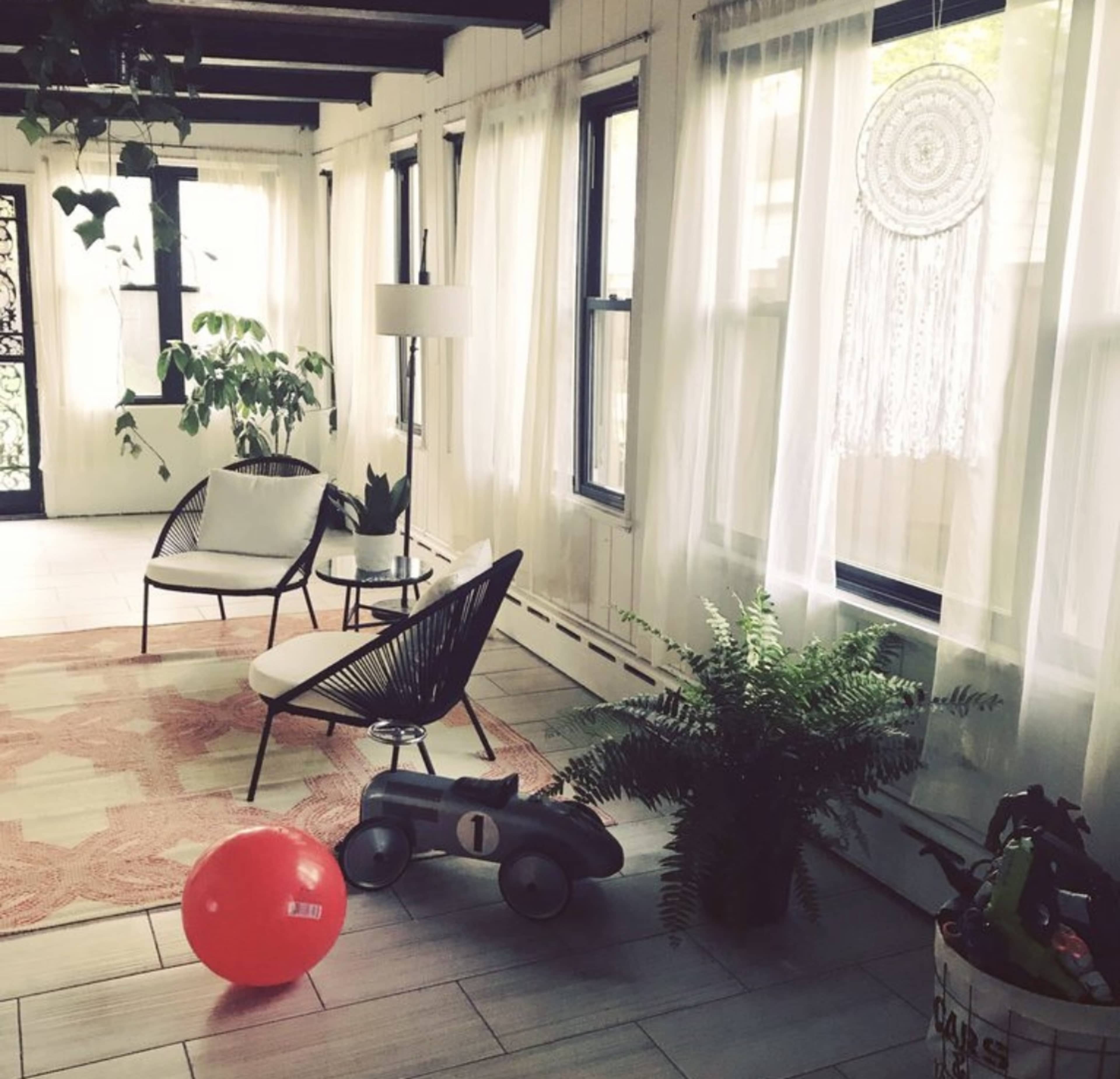 A bright room features two chairs, a lamp, a dreamcatcher hanging by the window, a potted fern, a red ball, and a toy car on a patterned rug.