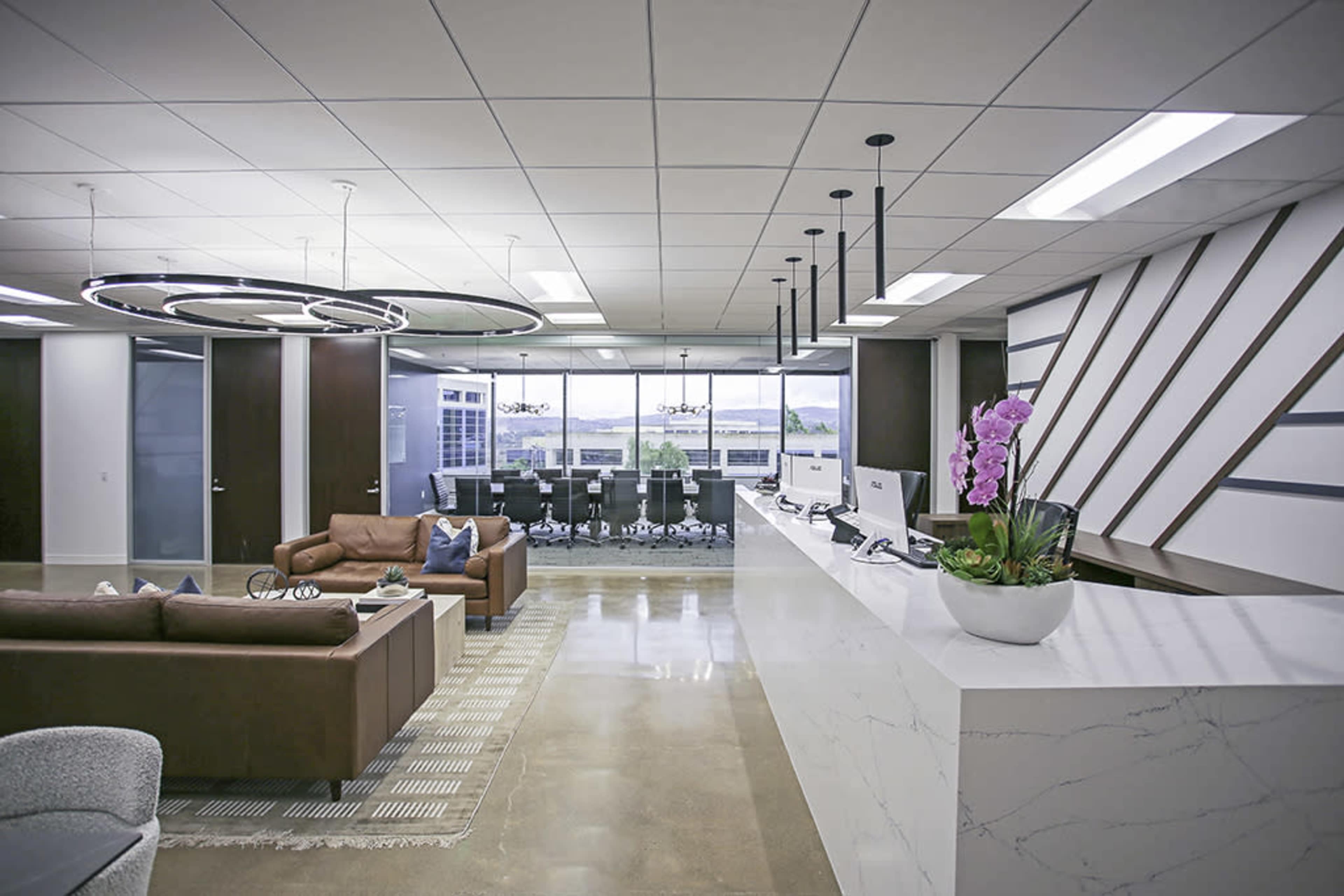 The image shows a modern office reception area with a white marble counter, a seating area with a brown couch, and a large glass meeting room in the background.