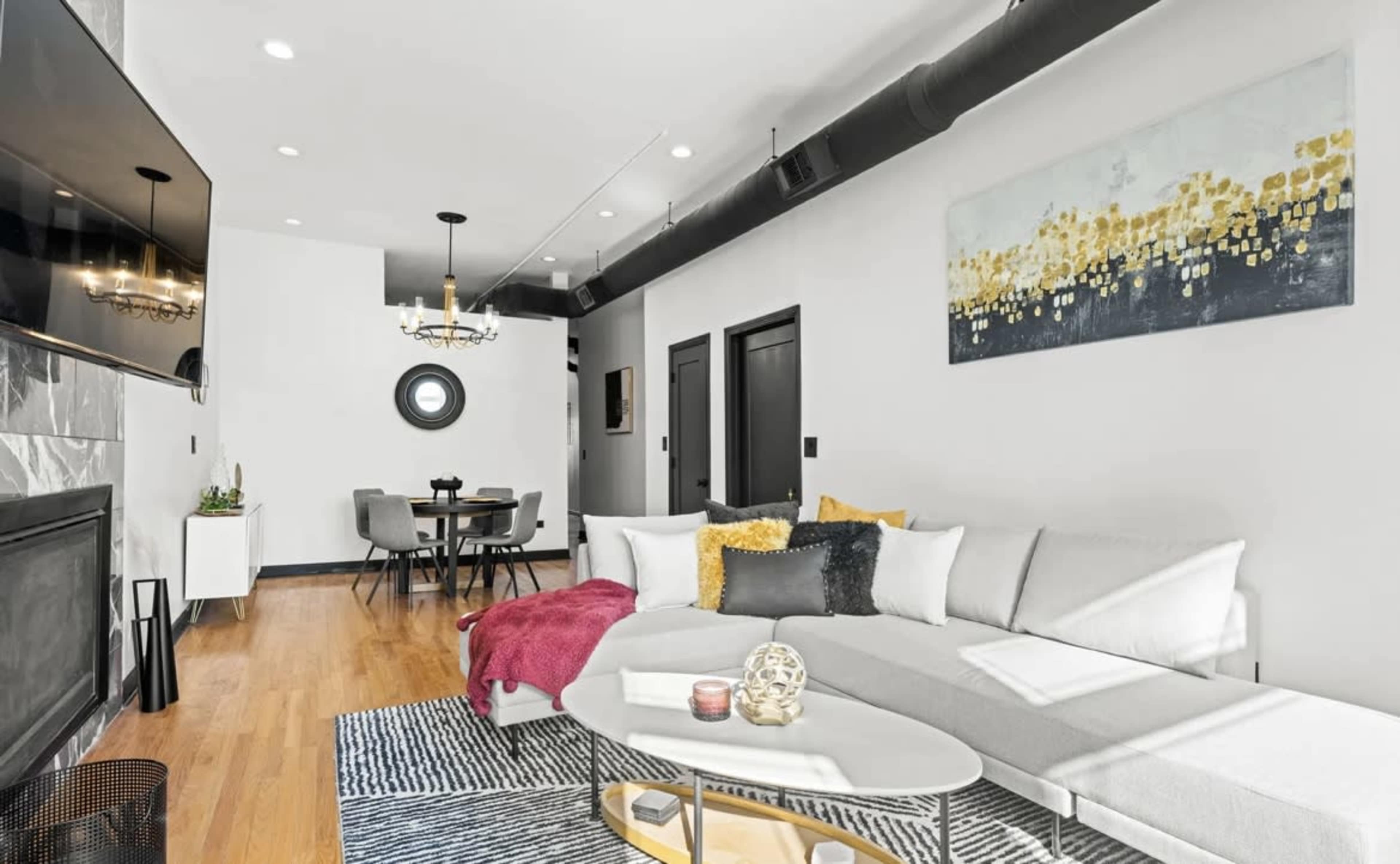 A modern living room with a gray sectional sofa, a large flat-screen TV mounted on the wall, and a round dining table in the background.