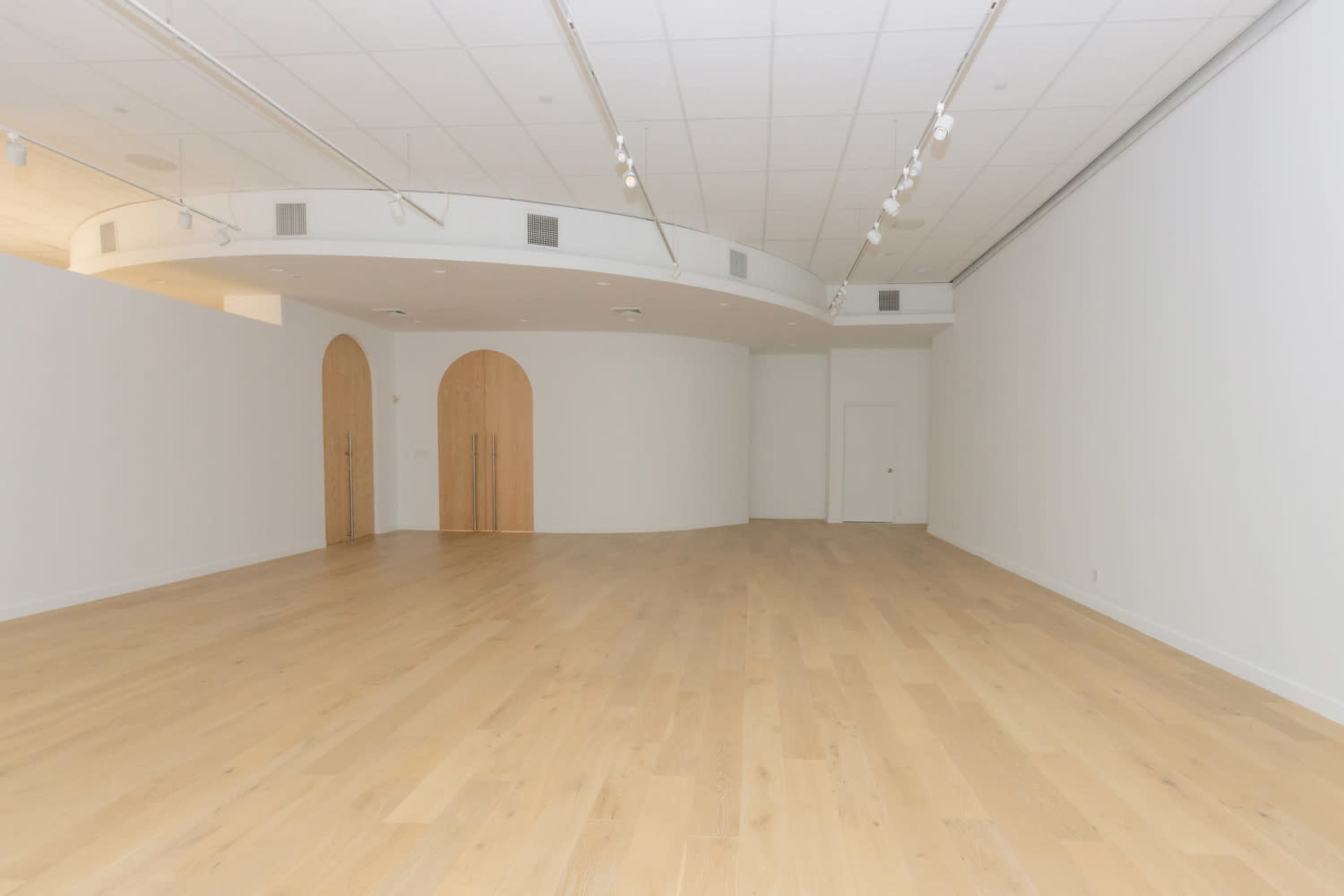 The image shows a spacious, empty gallery with light wooden flooring and curved walls, featuring two arched doorways.