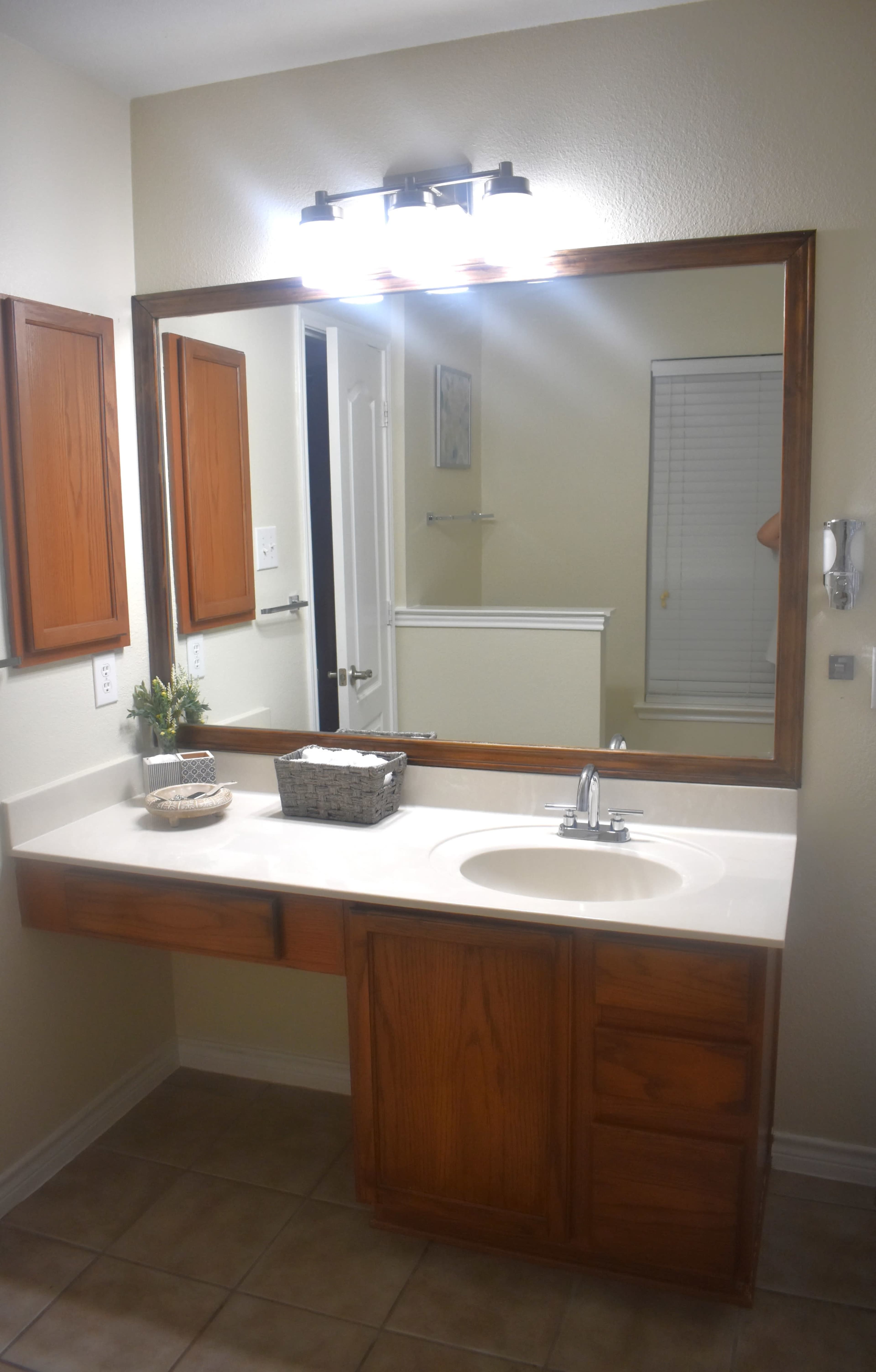 The image shows a bathroom with a wooden vanity, a large framed mirror above it, and a light fixture mounted on the wall.