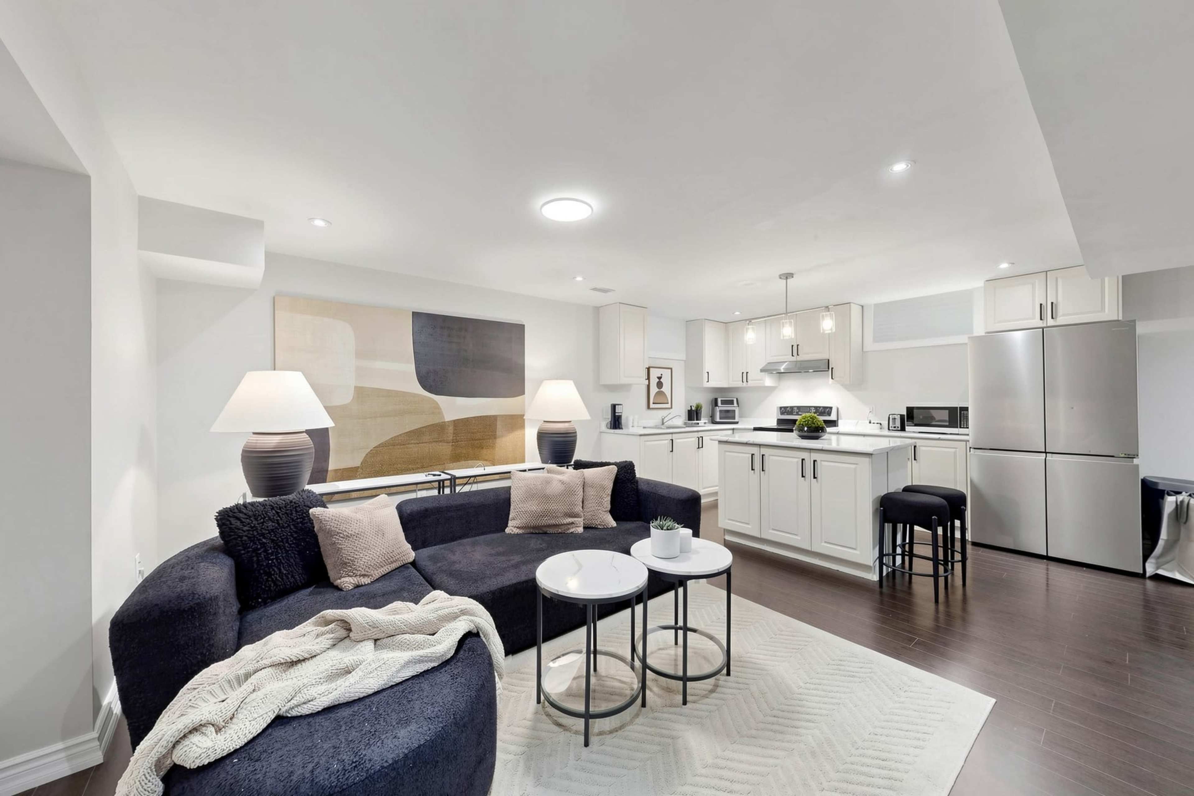 The image shows a modern living space featuring a dark blue curved sofa, two small round tables, and a kitchen area with stainless steel appliances.