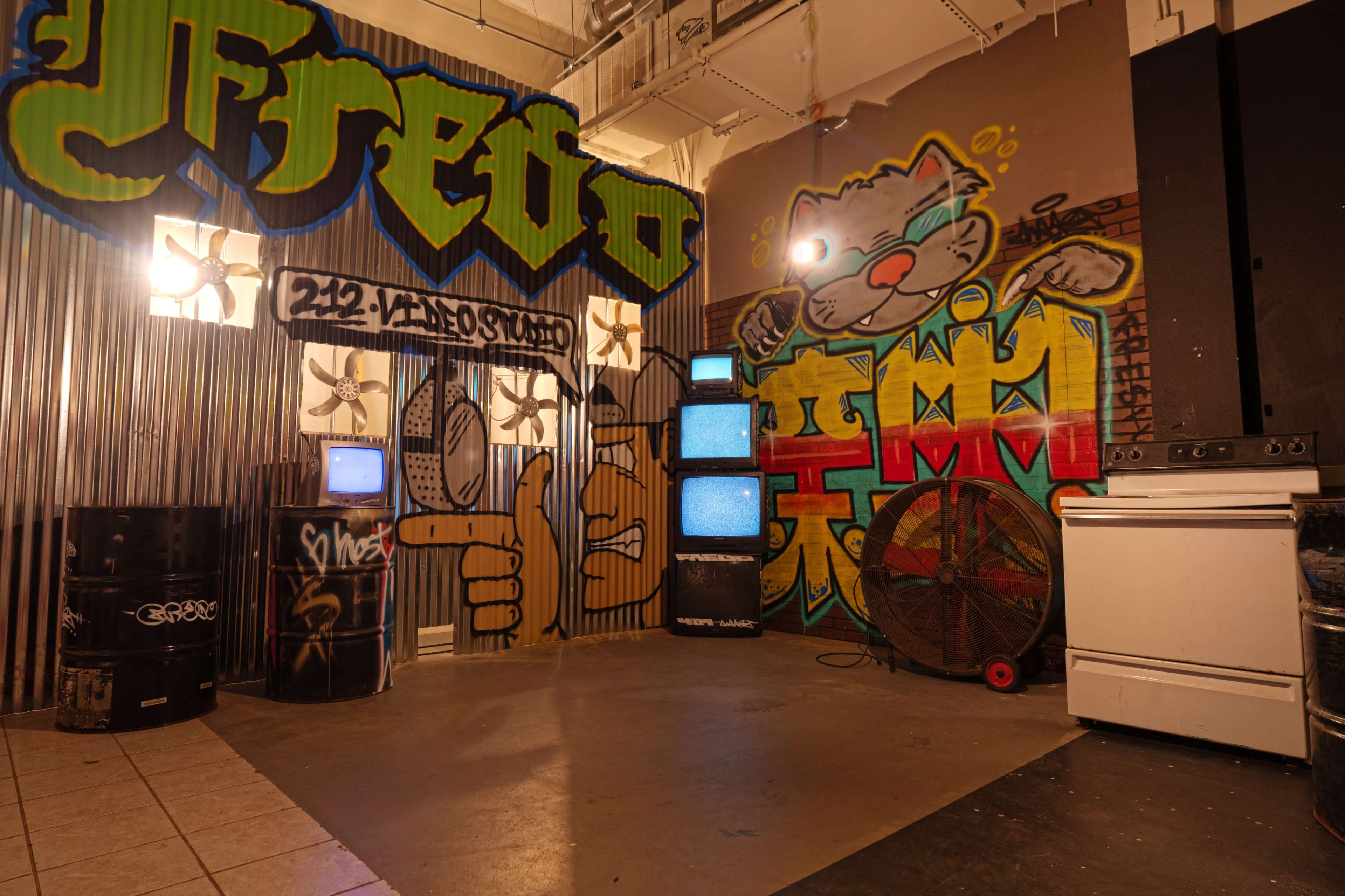 The image shows an indoor space decorated with colorful graffiti, featuring old televisions on a display stand, a large fan, and various industrial materials.