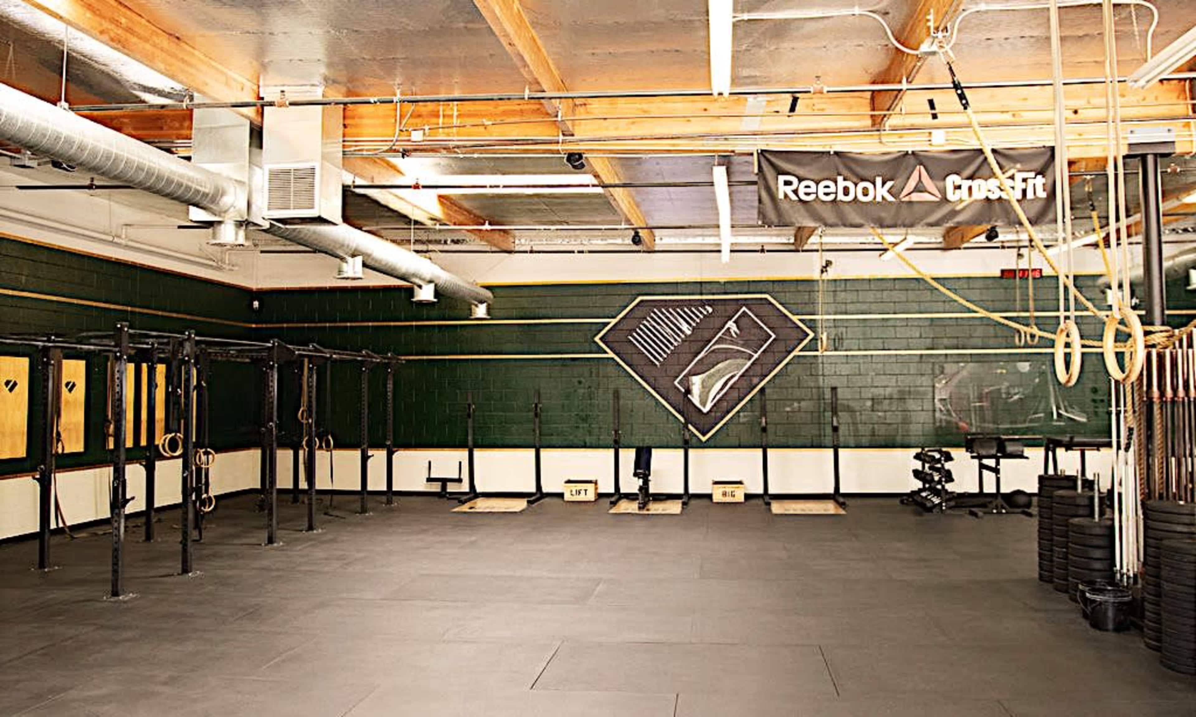 A spacious gym area featuring a set of pull-up bars, weightlifting platforms, and fitness equipment, with a Reebok CrossFit banner displayed on the wall.