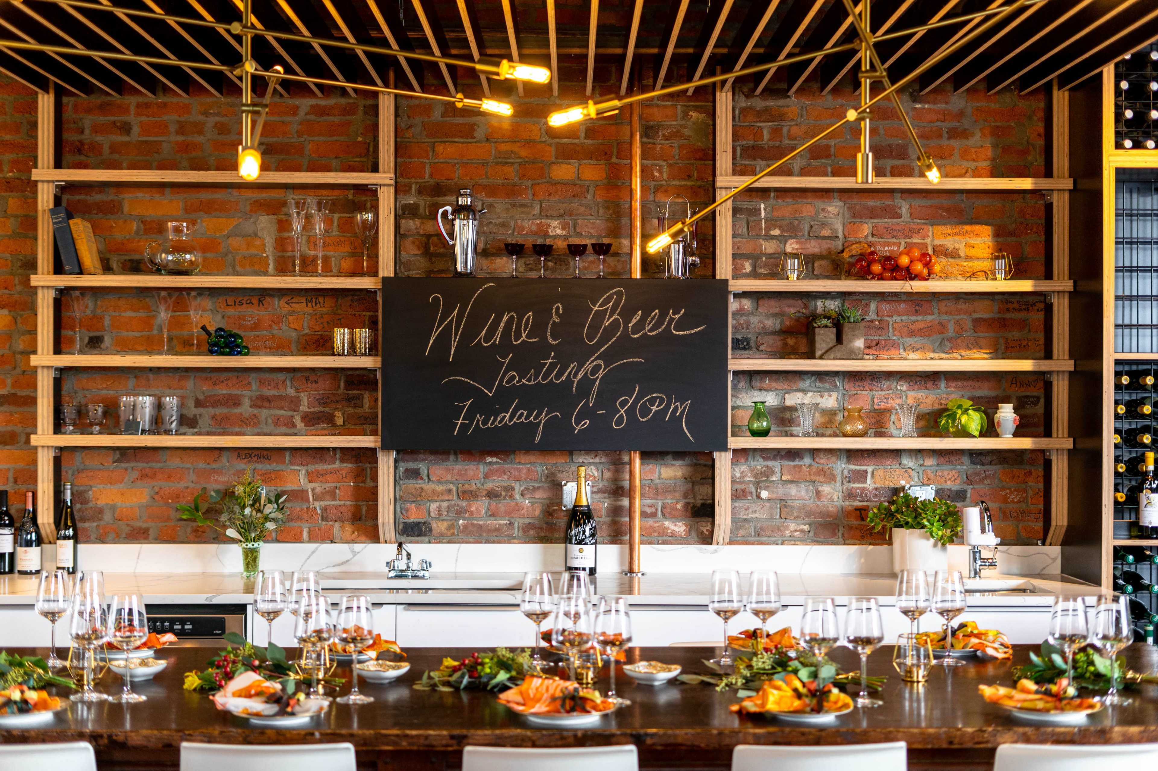 A long dining table is set with food and wine glasses, facing a brick wall with a chalkboard announcing a wine and beer tasting event.