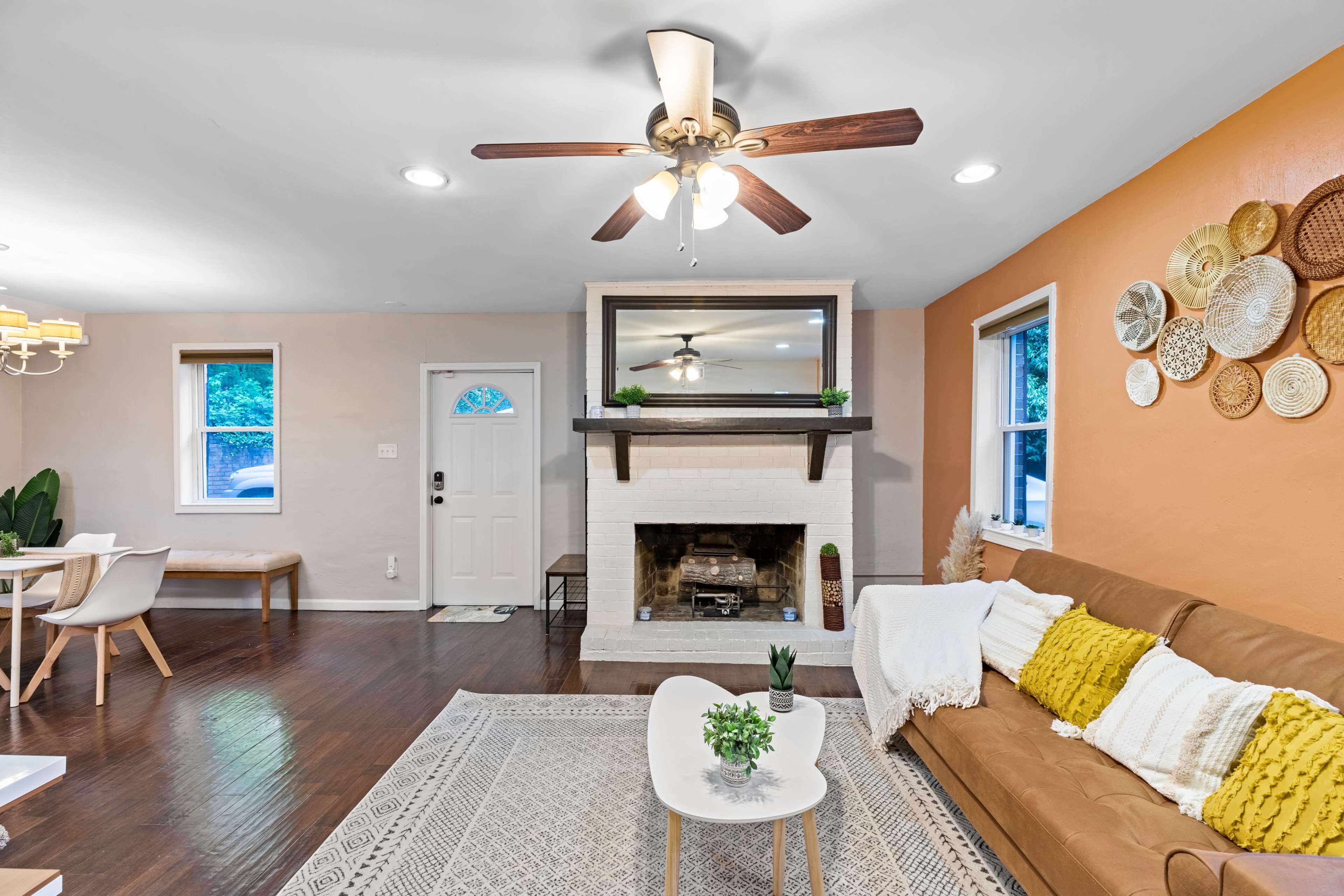 The image shows a living room with a brown couch, a fireplace with a mirror above it, and a dining area with a light-colored table and chairs.