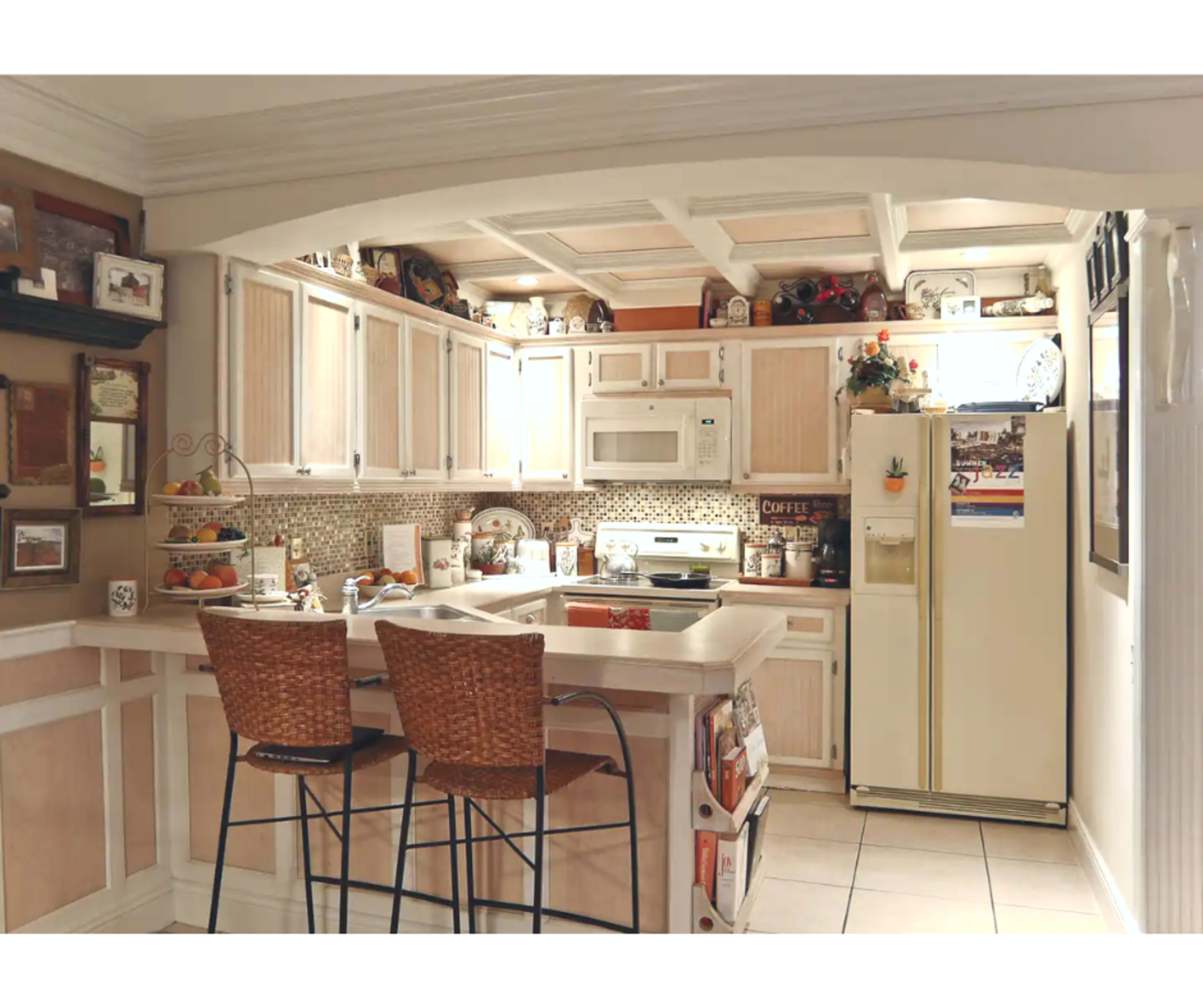 A kitchen features wooden cabinetry, a countertop with bar stools, and a refrigerator, with various decor and appliances arranged throughout the space.