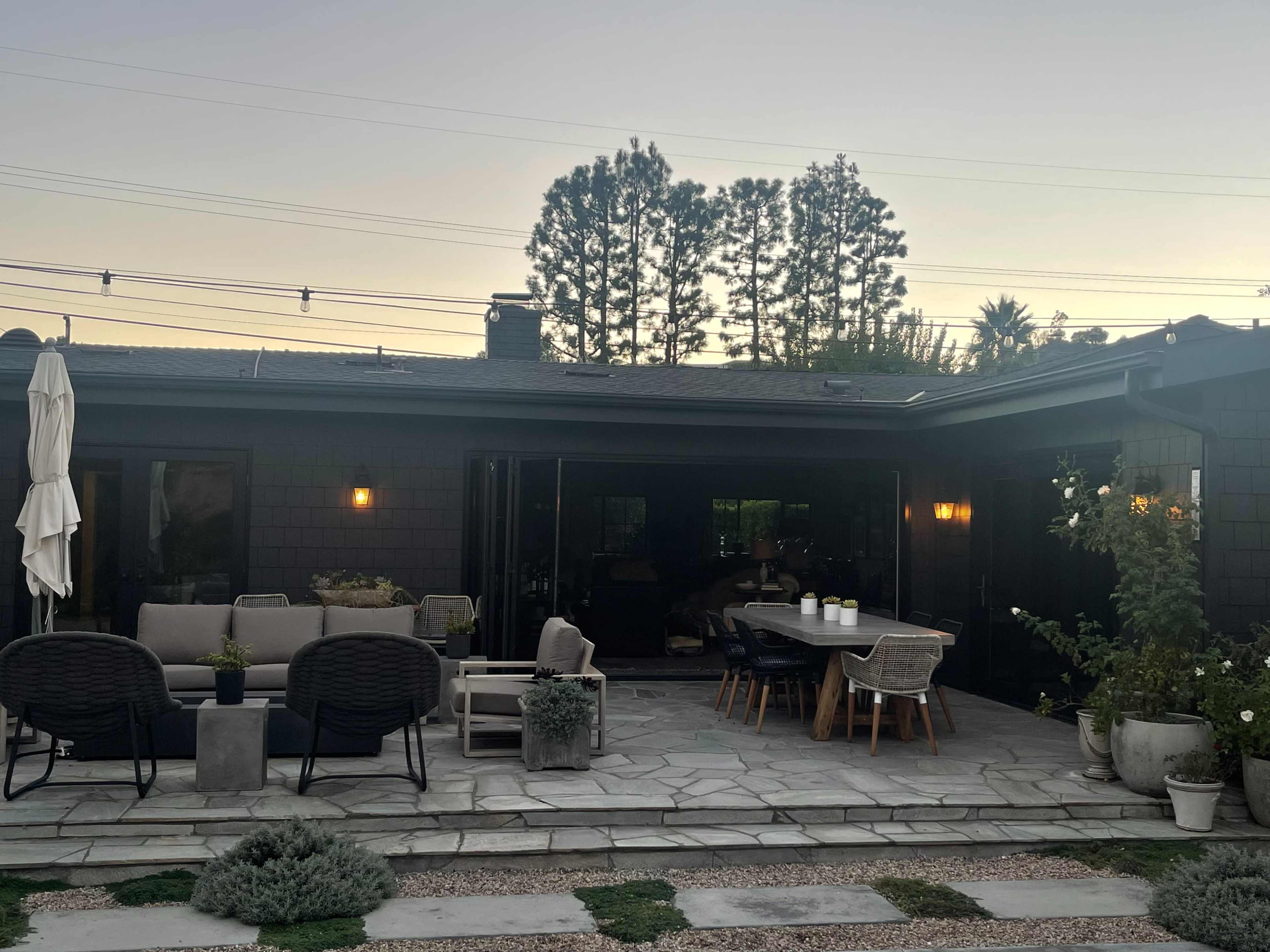 The image shows a modern outdoor patio area with seating arrangements and a dining table, surrounded by plants and illuminated by wall sconces.