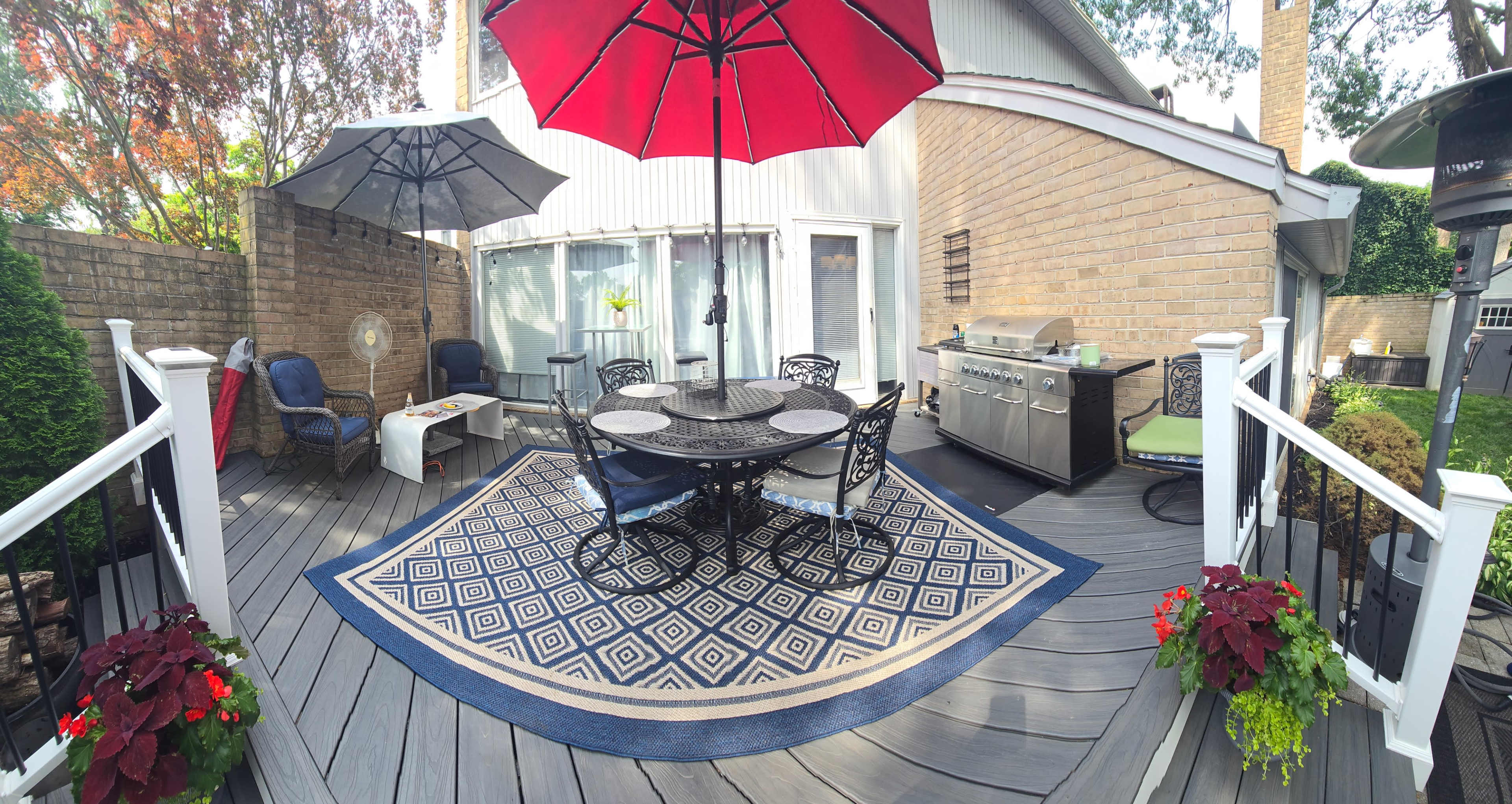 The image shows a patio area with a round dining table and six chairs, a red umbrella, a grill, and decorative plants, all set on a wooden deck.