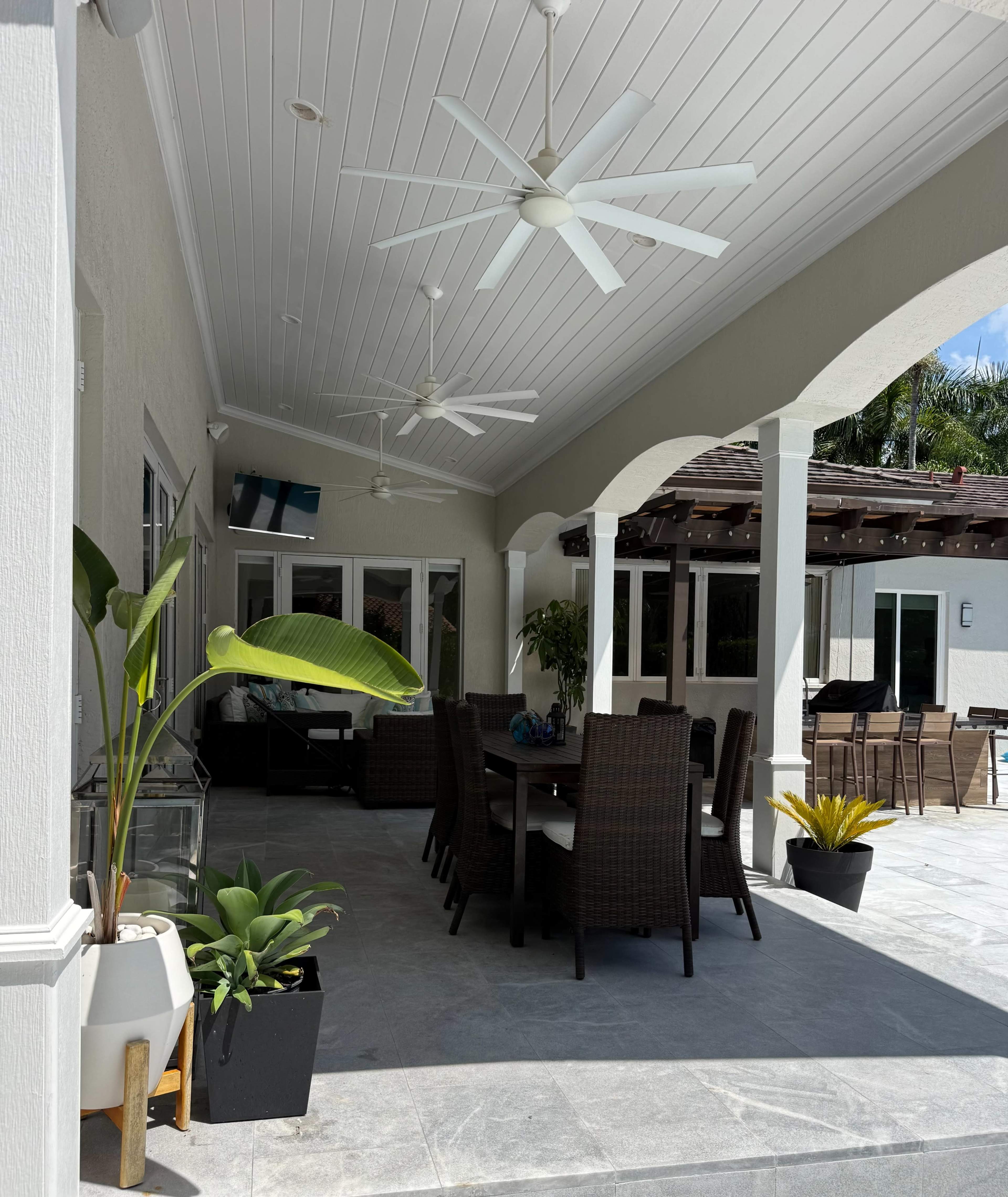 The image shows a covered outdoor patio with a dining table, chairs, and ceiling fans, surrounded by potted plants.
