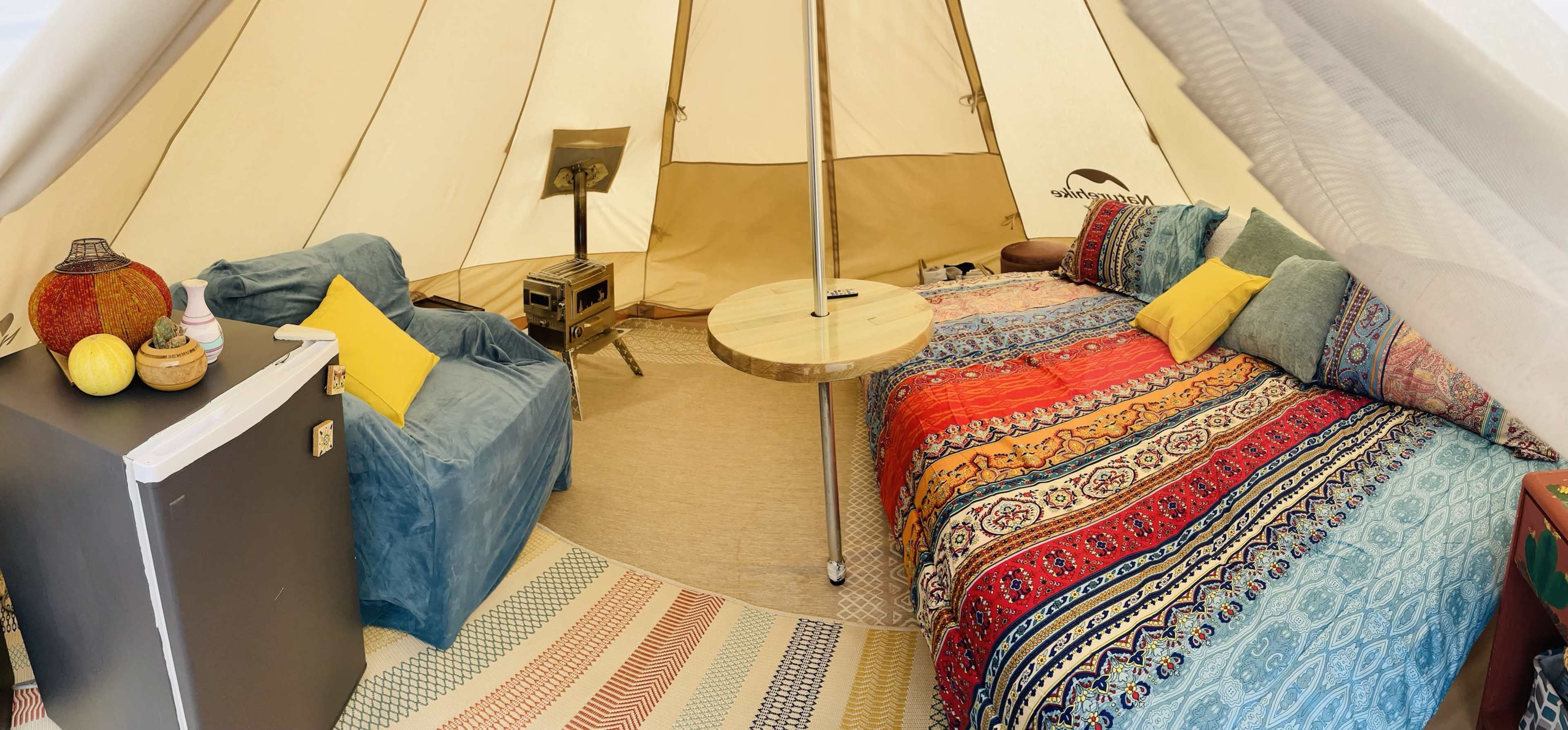 The interior of a large tent features a couch, a circular wooden table, a bed with colorful bedding, and a small refrigerator.