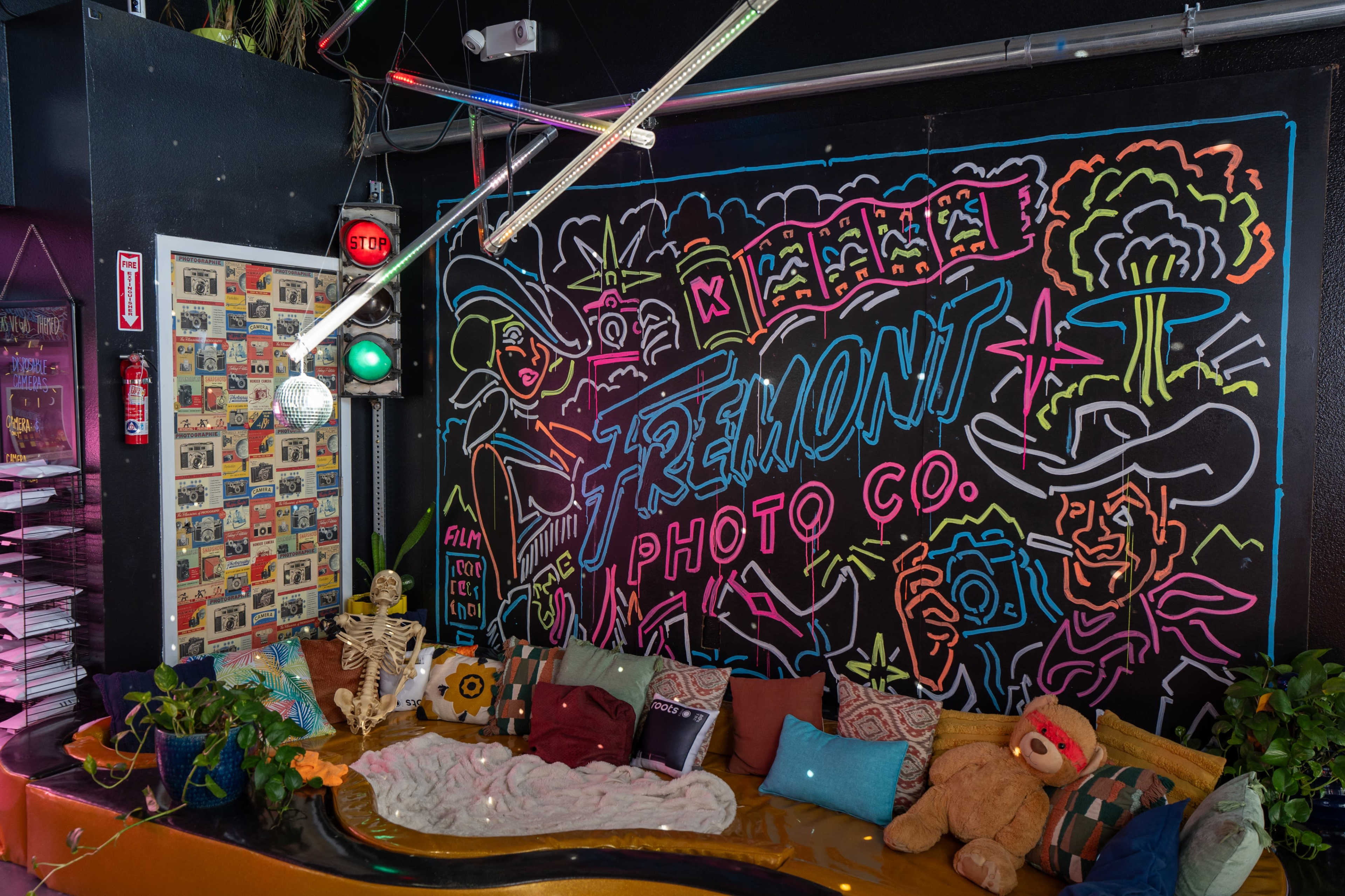 The scene features a colorful neon mural of cowboys and cameras on a black wall, with a cozy seating area adorned with pillows, plants, and whimsical decor.