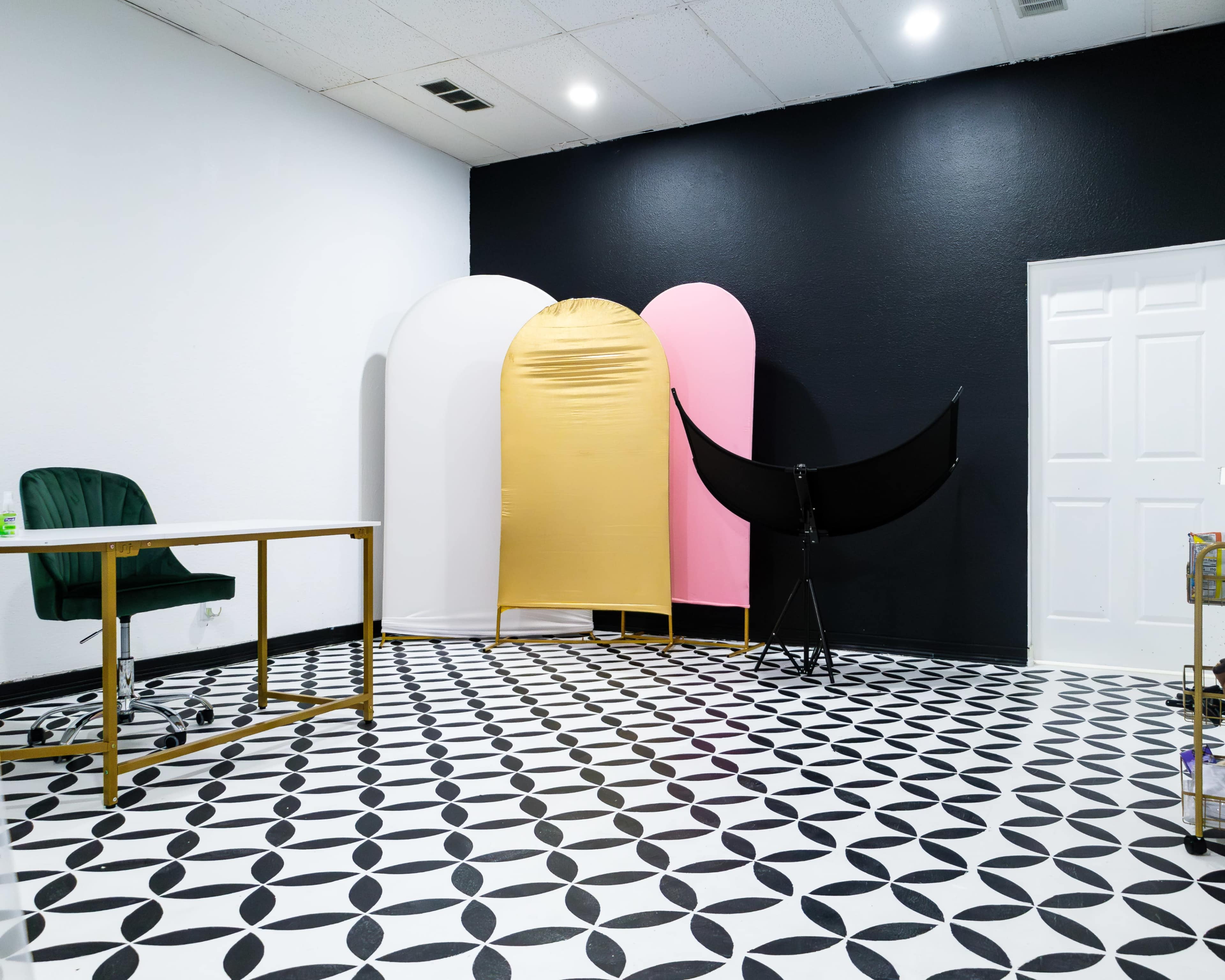 A modern studio space with patterned black and white tile flooring, a gold, pink, and white backdrop setup, and a folding screen, along with a green chair and a table.