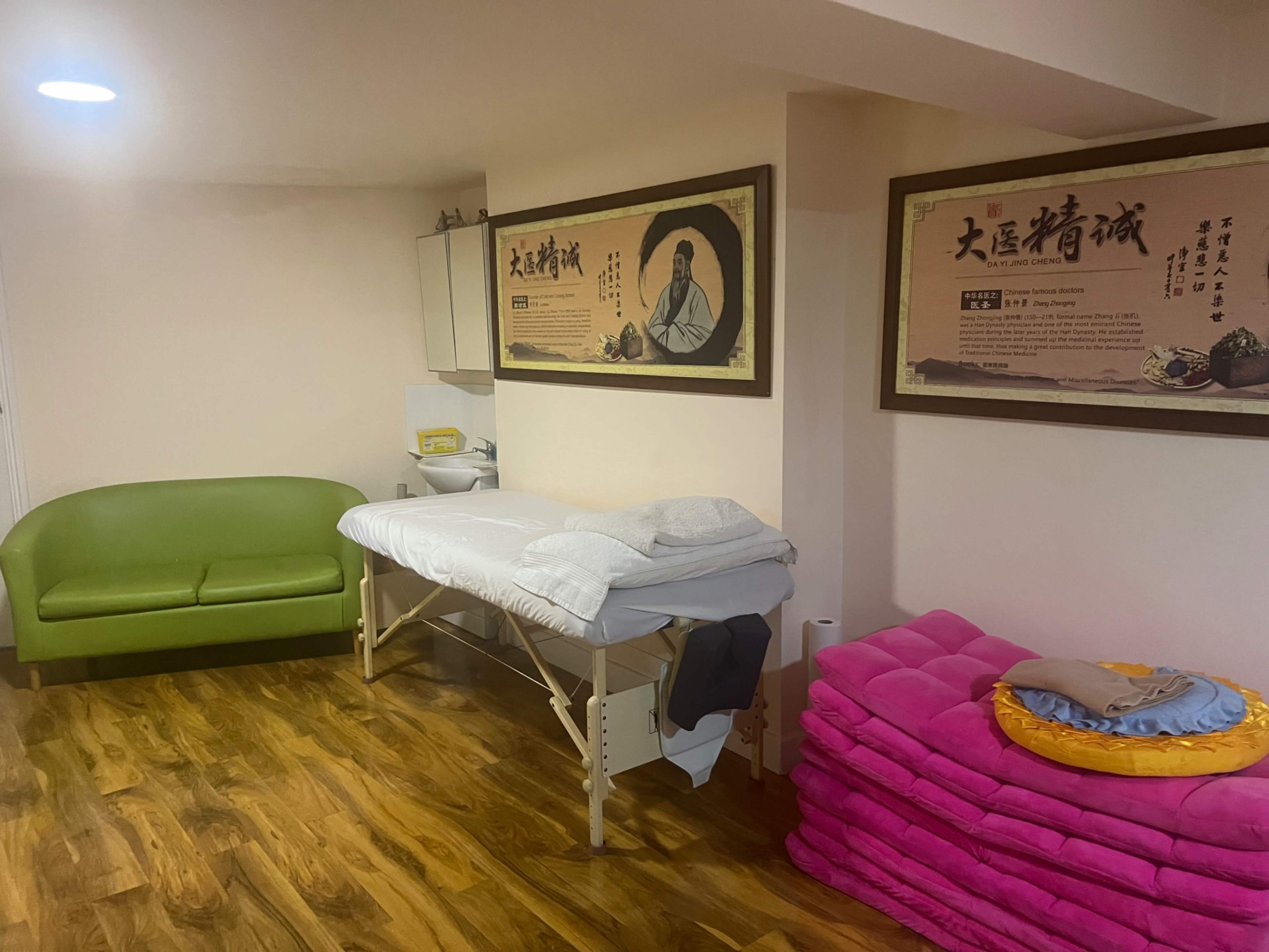 The image shows a massage therapy room with a green couch, a treatment table covered in white linens, and a stack of pink cushions on the floor, along with framed traditional artworks on the walls.