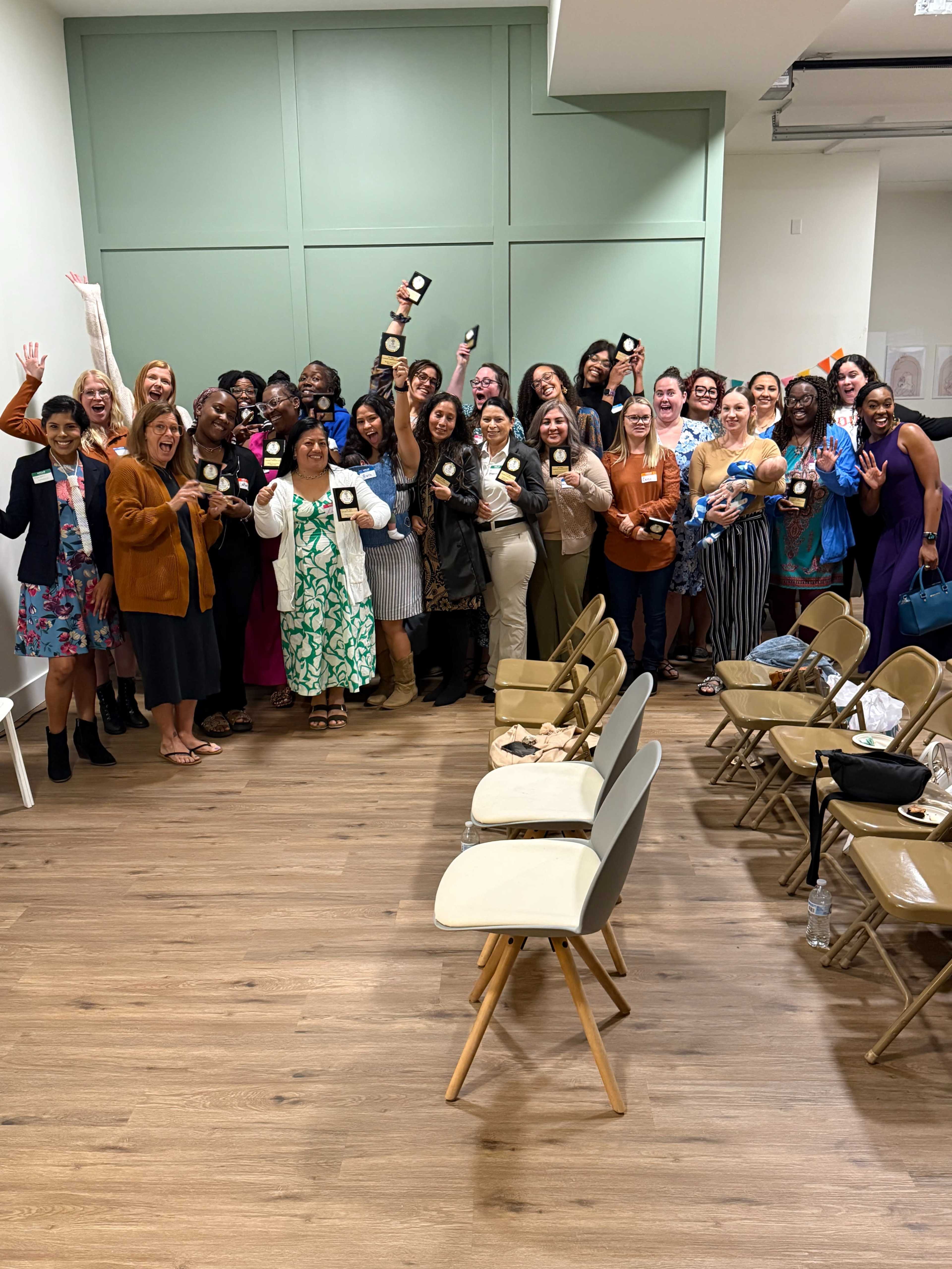 A large group of women poses together in a room, many holding awards and celebrating, with a few empty chairs in the foreground.