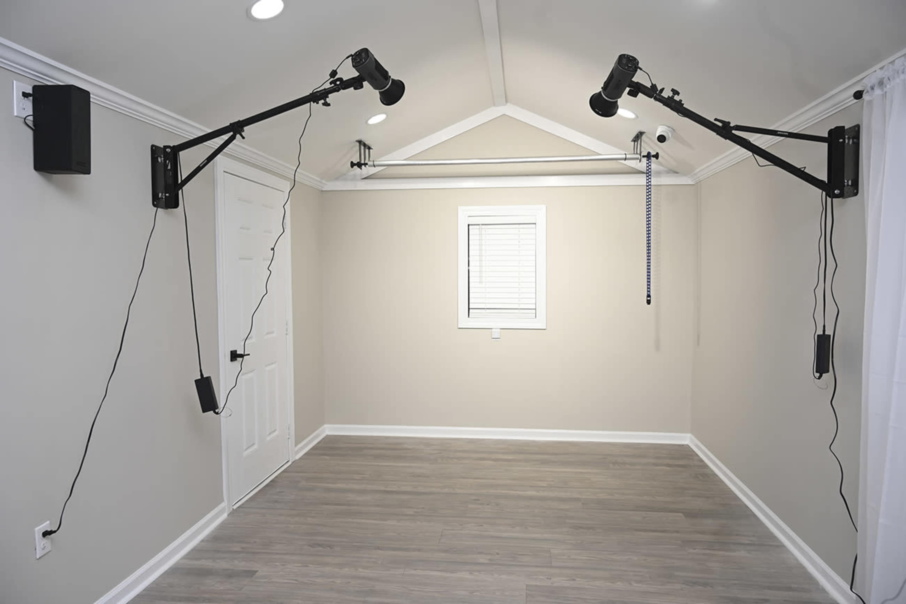 The image shows a sparsely furnished room with light wooden flooring, a white door, a small window with blinds, and two adjustable mounting brackets for lighting.
