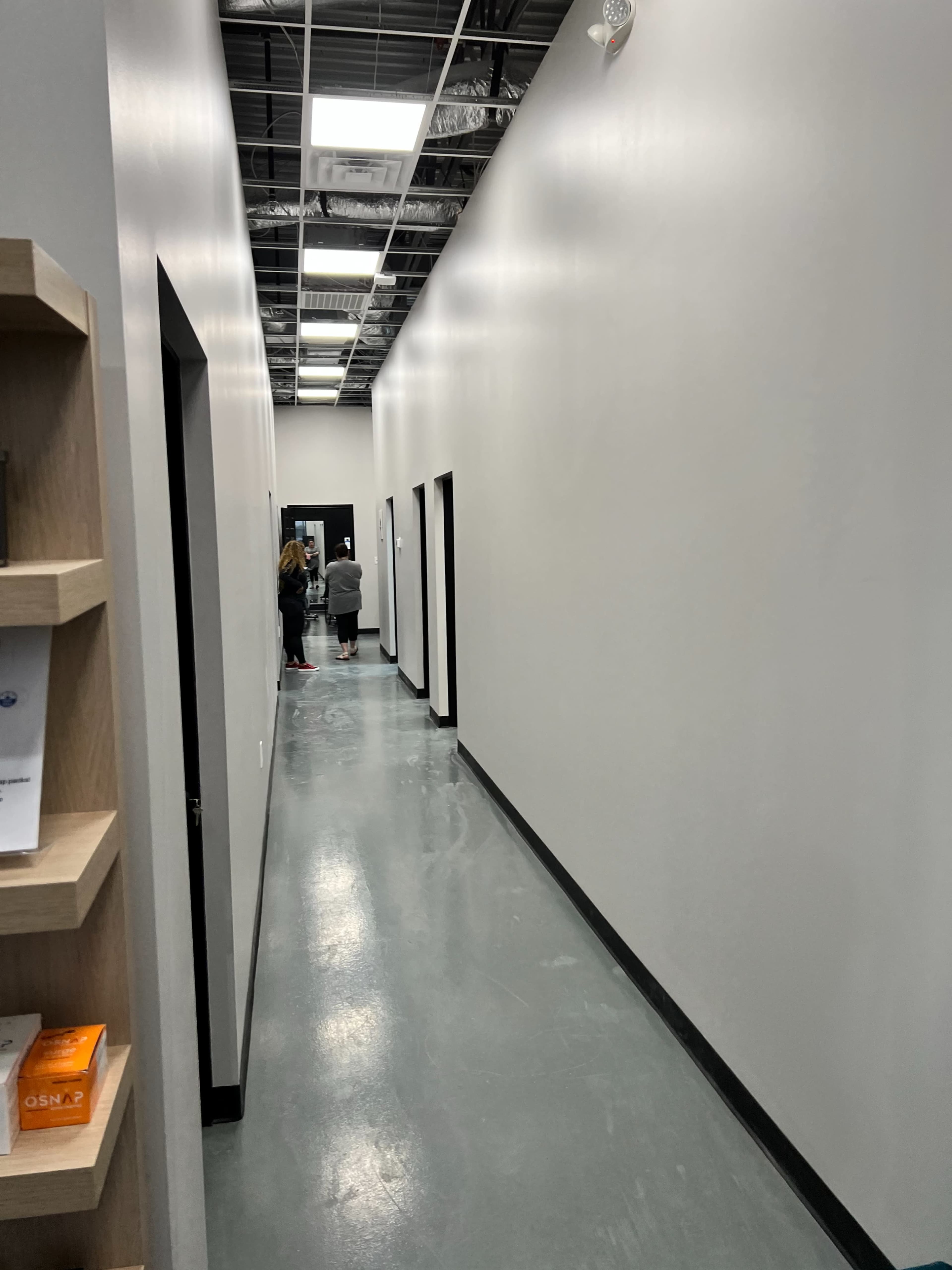 A narrow hallway with gray walls and a concrete floor leads to open doors at both ends, where two people are visible.