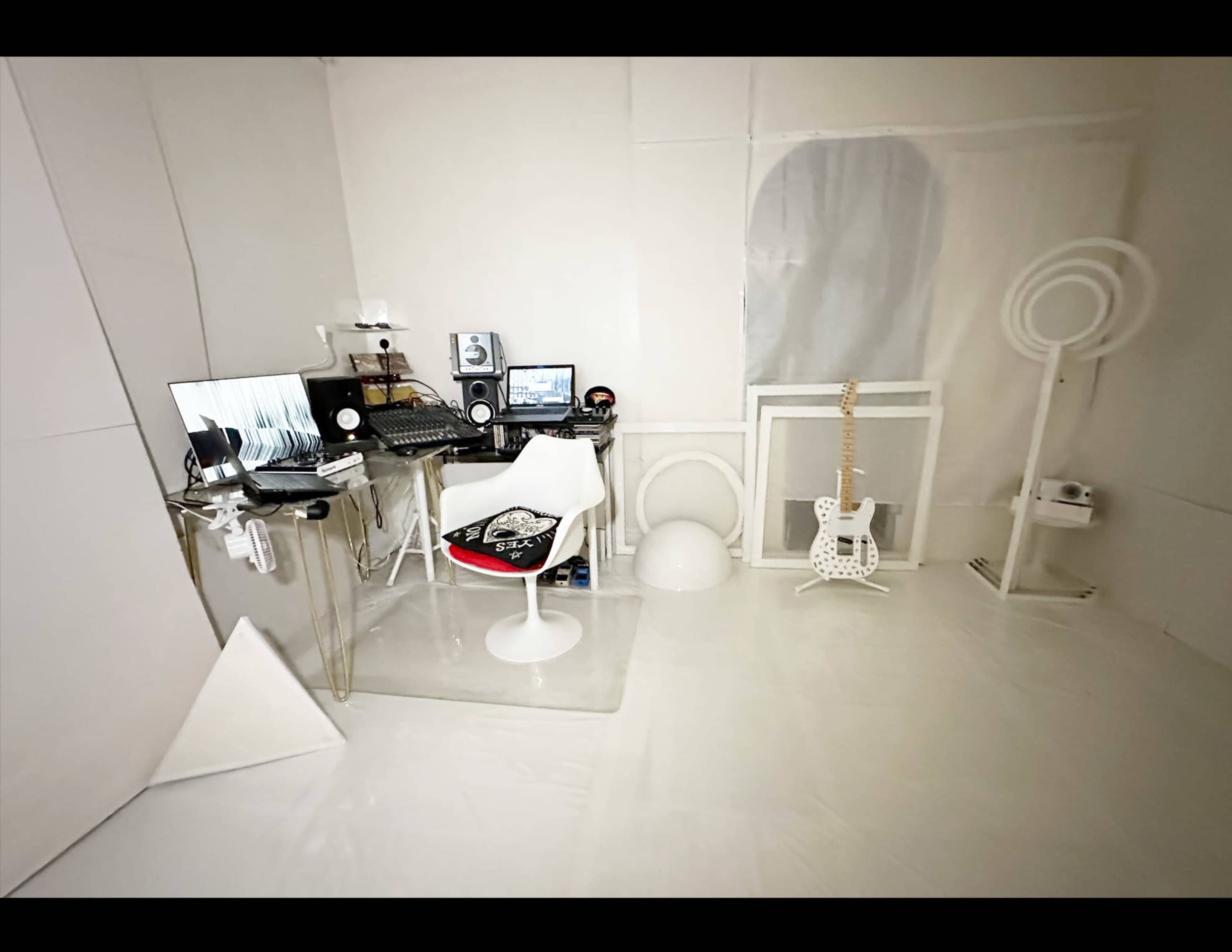 The image shows a minimalist white studio space with a desk, computer equipment, a guitar, and various geometric objects.