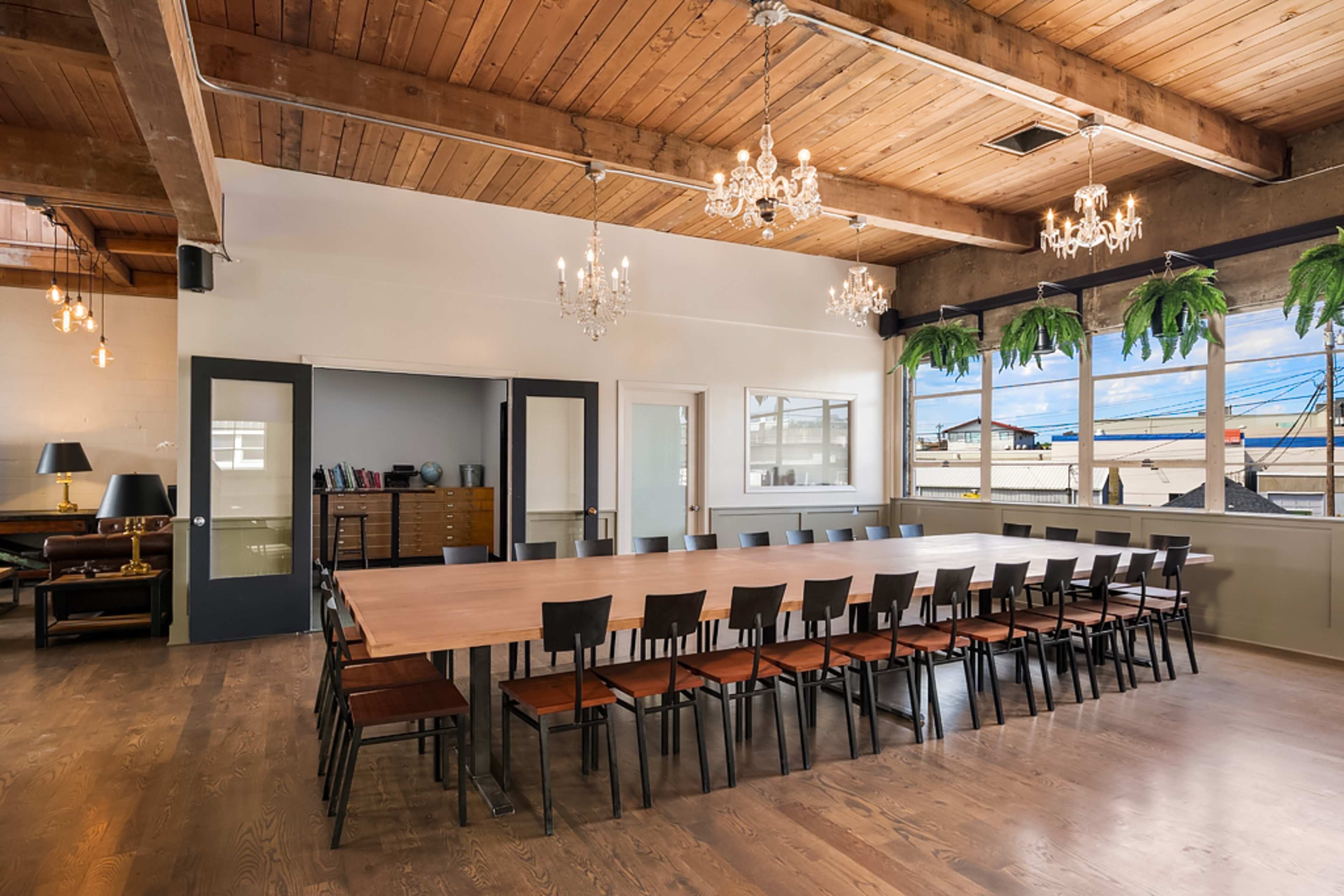 A spacious meeting room with a long wooden table surrounded by black chairs, featuring large windows and a wooden ceiling with chandeliers.