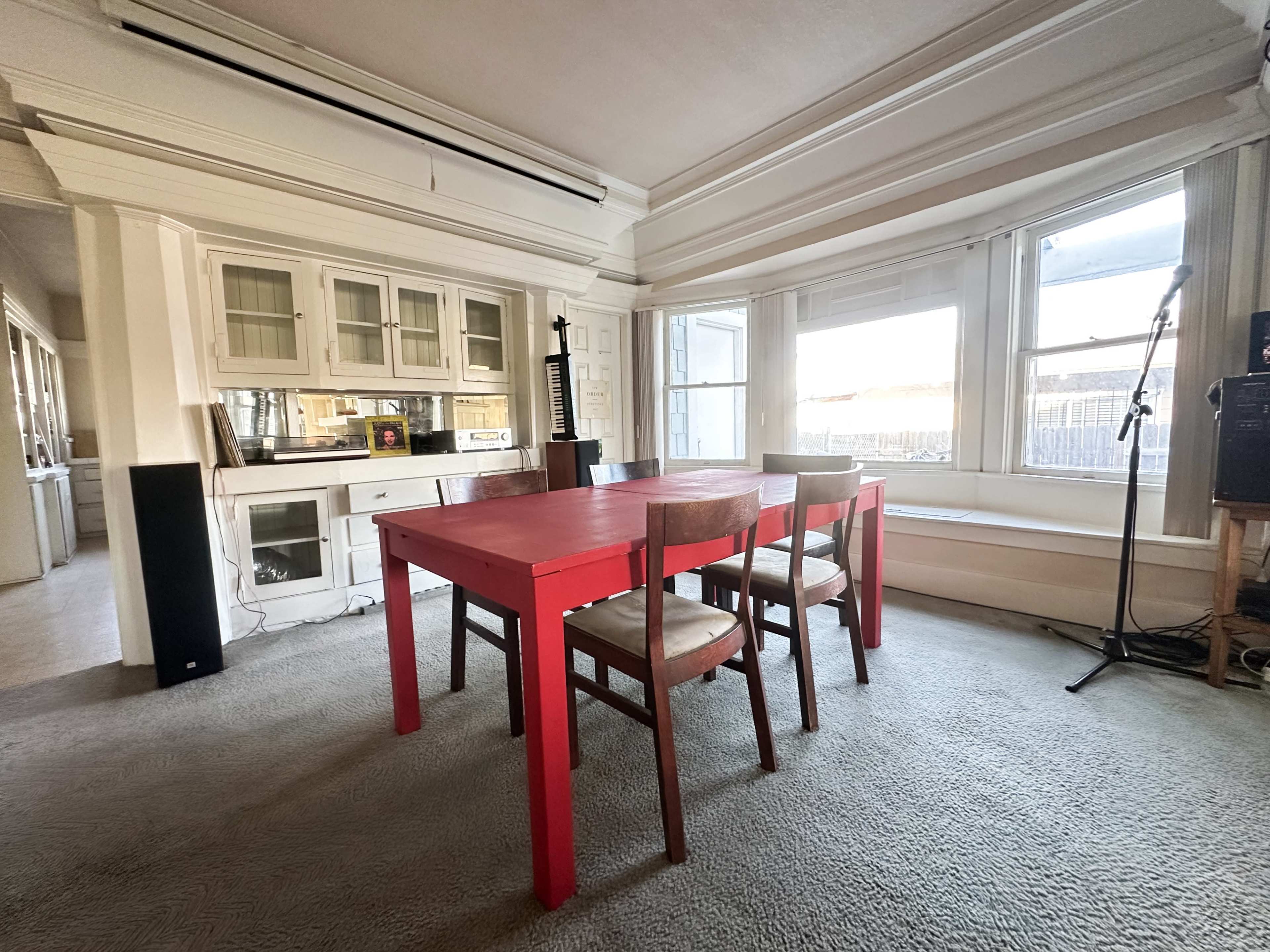 The image shows a spacious dining area with a red table and wooden chairs, situated near large windows that provide natural light.