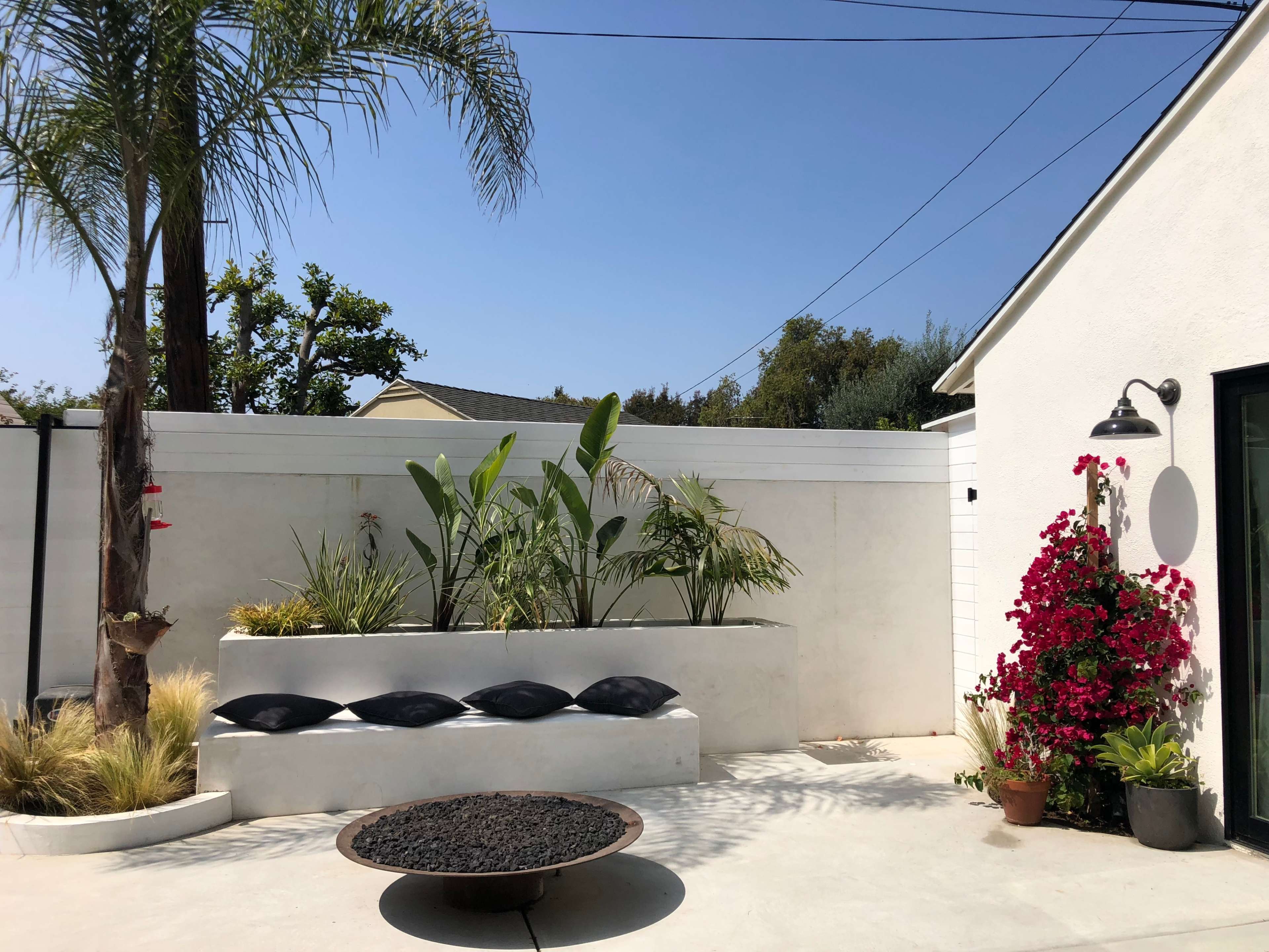 A modern outdoor space featuring a concrete planter with greenery, a round fire pit, and a wall adorned with vibrant flowers against a clear blue sky.