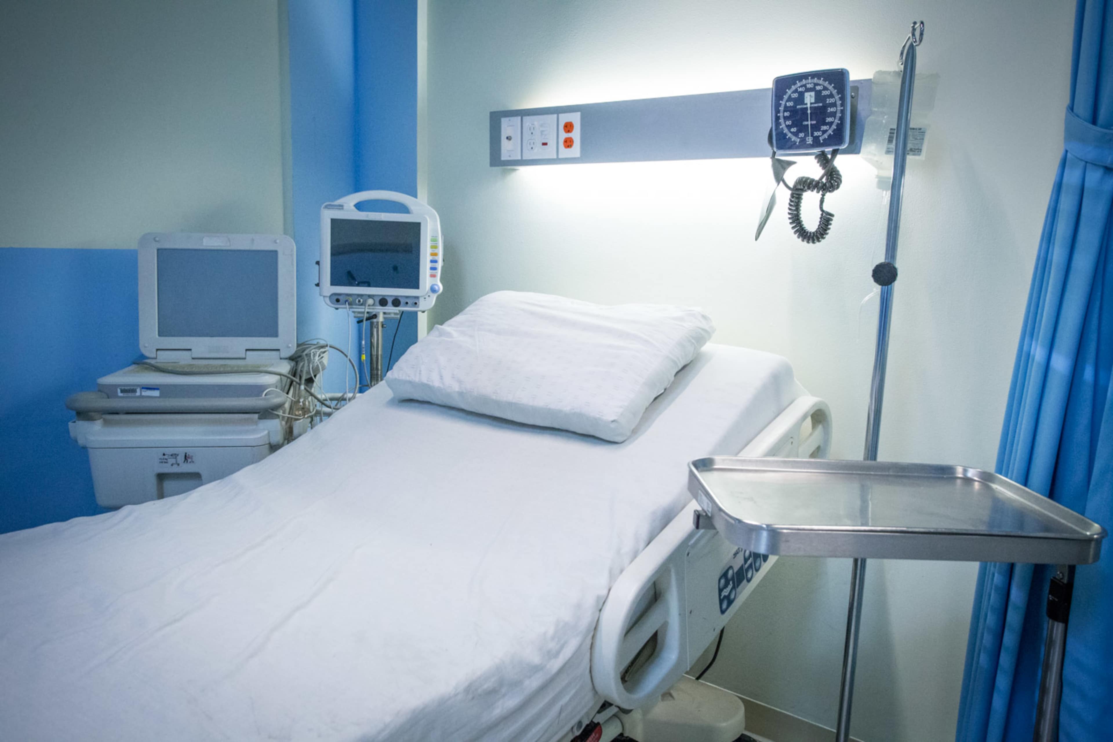 A hospital room with a vacancy, featuring a bed with white linens, a medical monitor, and a wheeled tray beside it.