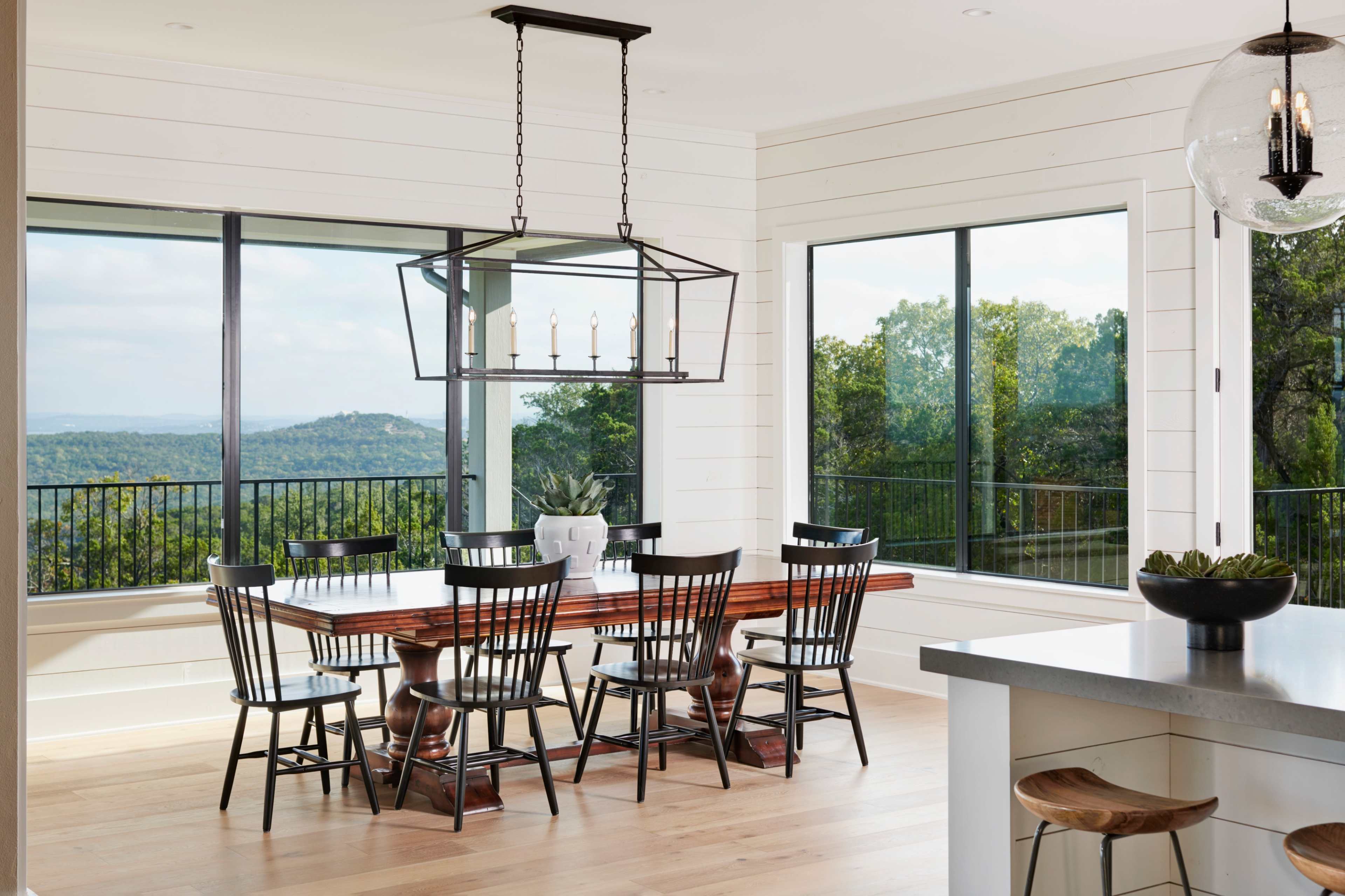 A spacious dining area features a long wooden table surrounded by black chairs, with large windows offering a view of the outdoors.