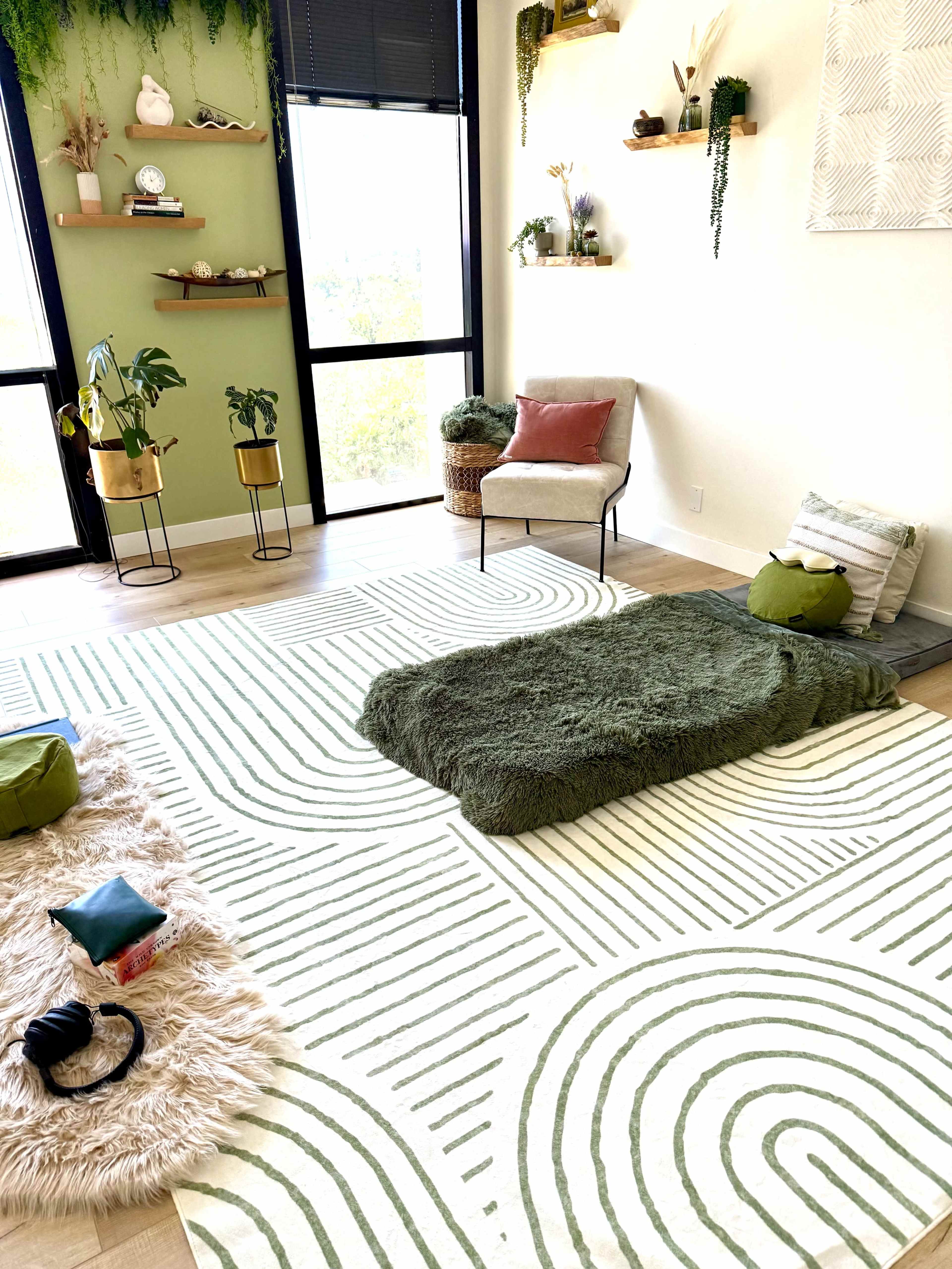 The image shows a modern interior space featuring a light-colored floor with patterned rugs, a few plants, a chair, and cushions arranged in a cozy seating area.