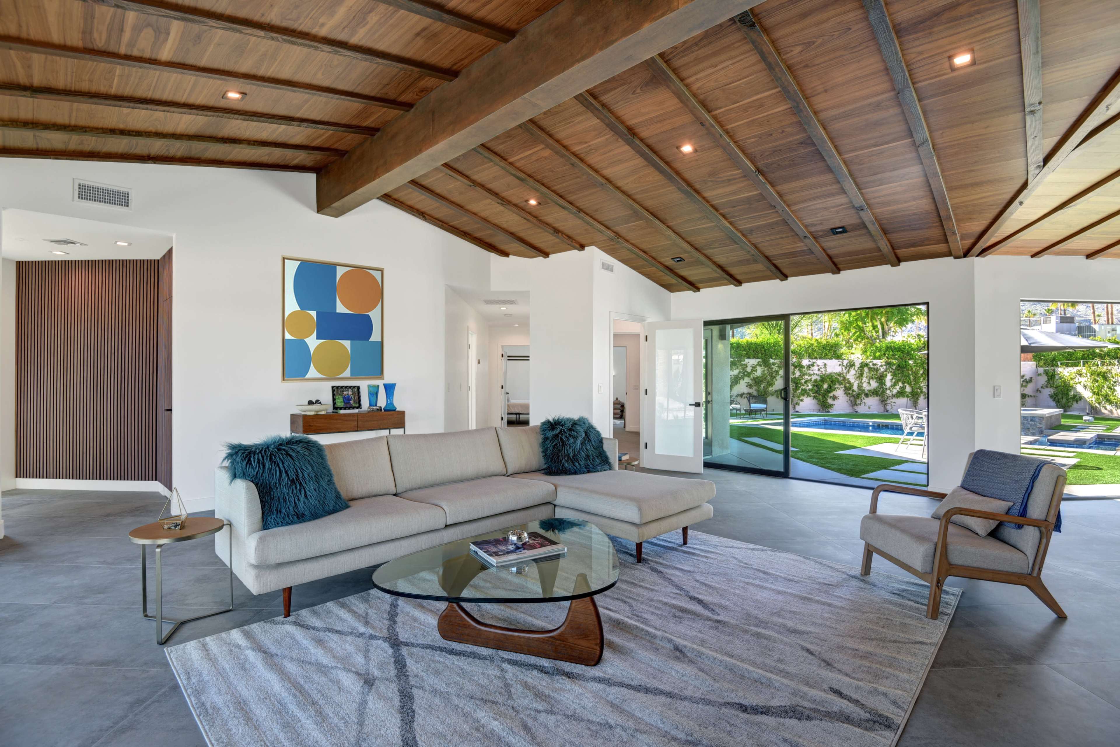 A modern living room with a sectional sofa, a glass coffee table, and large windows overlooking a grassy area and pool.