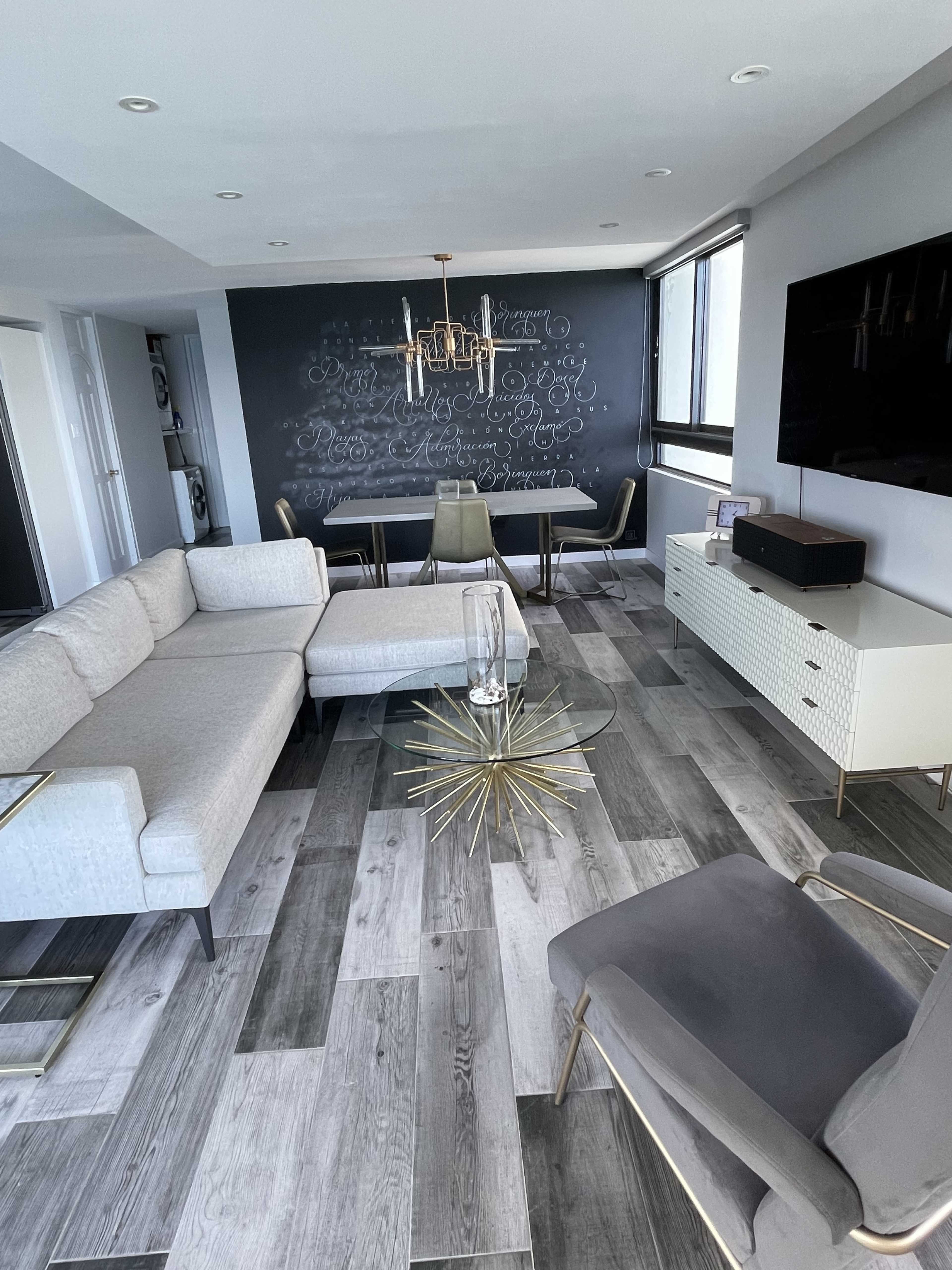 A modern living room features a light gray sectional sofa, a glass coffee table, a round dining table with chairs, and a dark chalkboard wall with decorative writing.