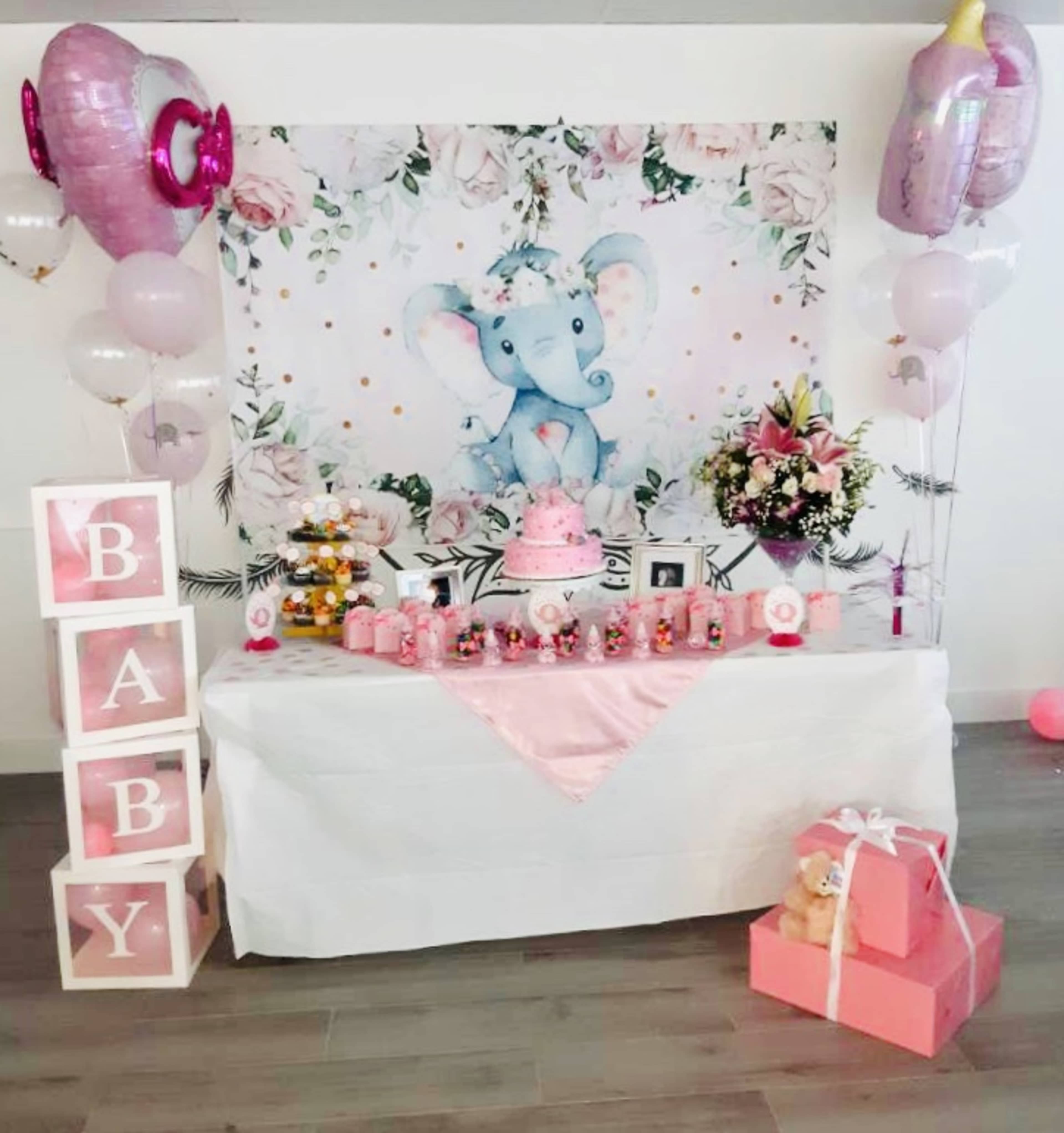 A decorated table features a pink-themed baby shower setup with an elephant backdrop, a cake, gifts, and floral arrangements.