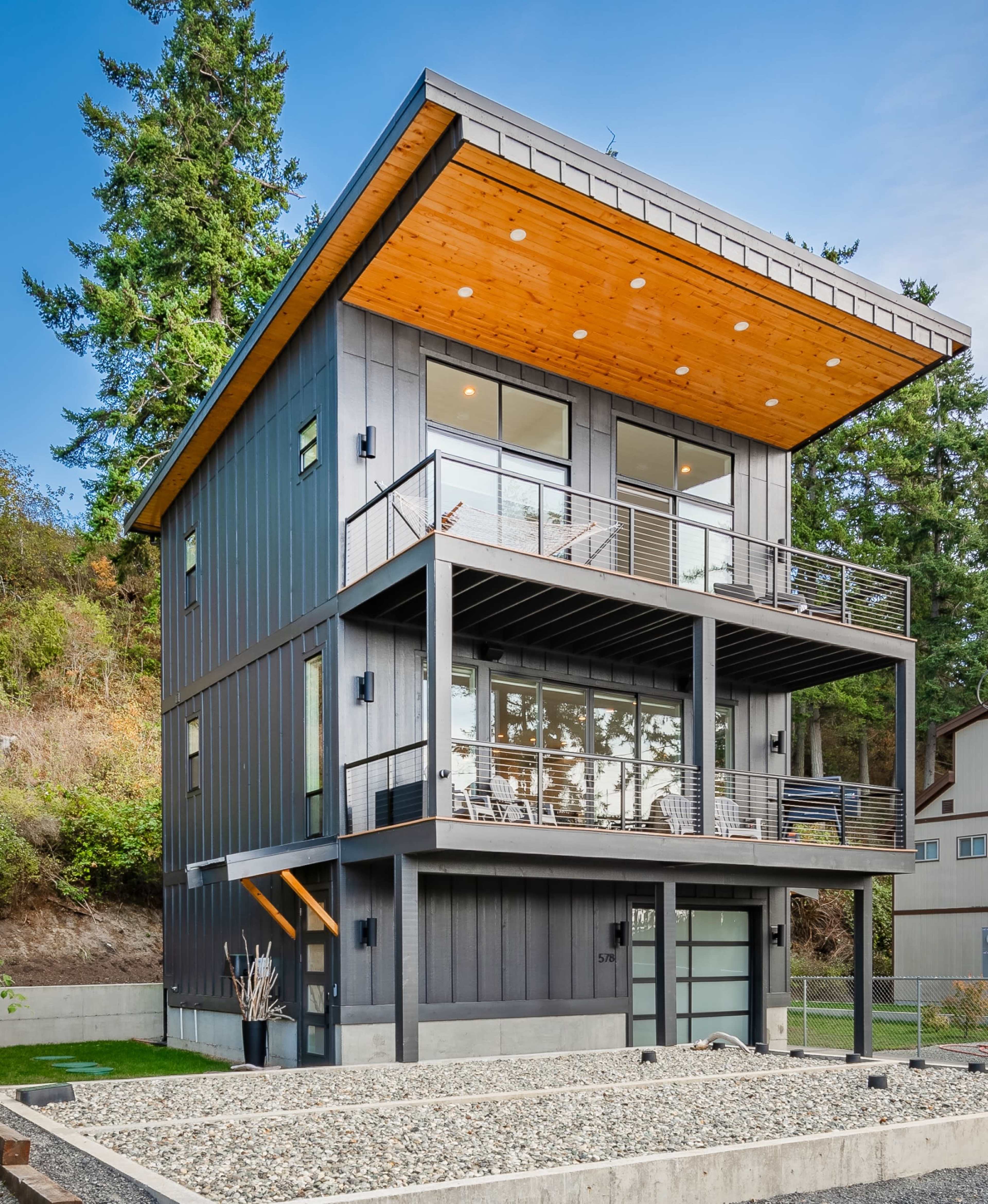 The image shows a modern two-story house with a wooden overhang, large windows, and a stone pathway.