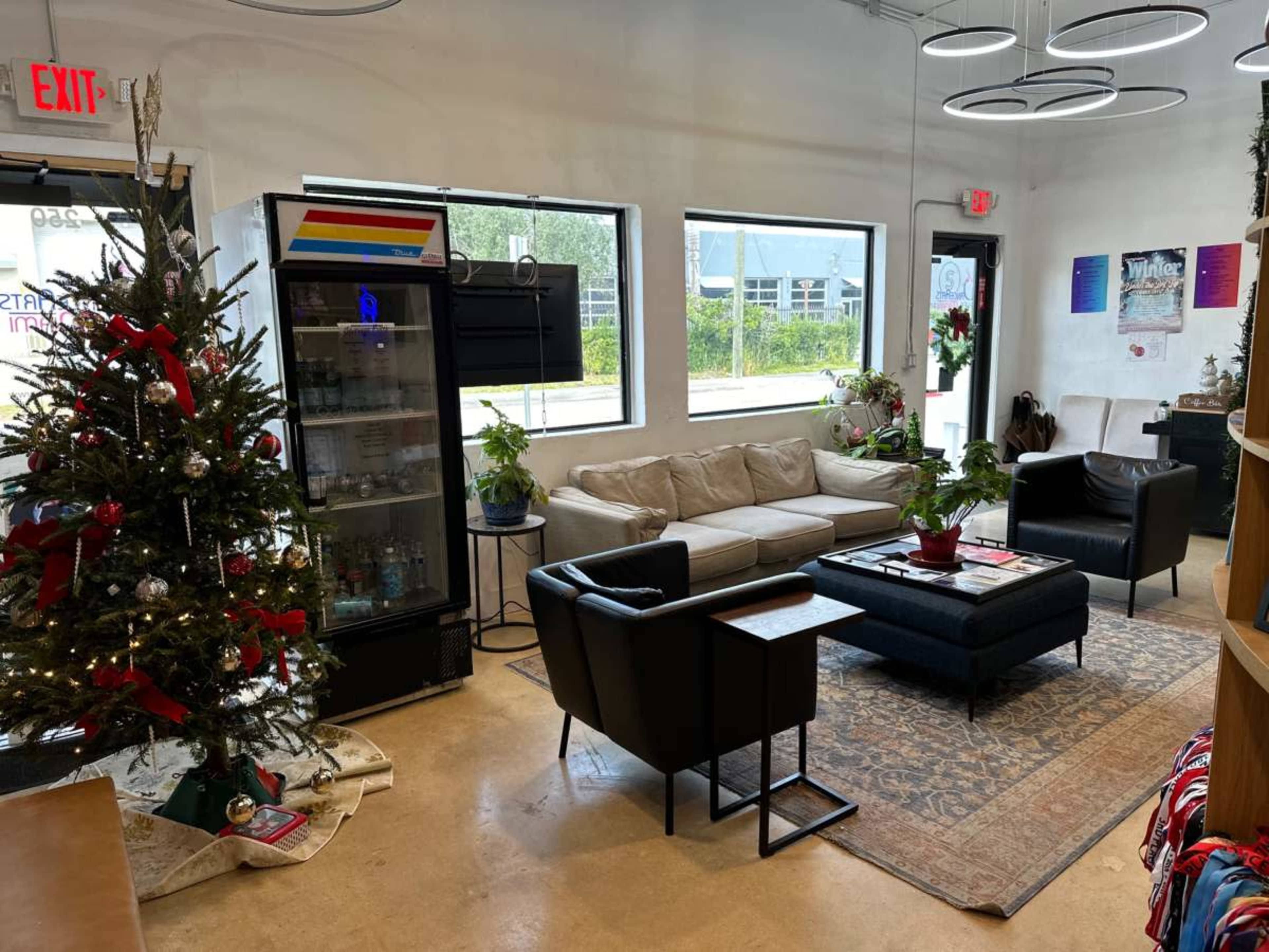 The image shows a cozy waiting area with a Christmas tree, a sofa, two armchairs, a coffee table, and a refrigerator displaying drinks.